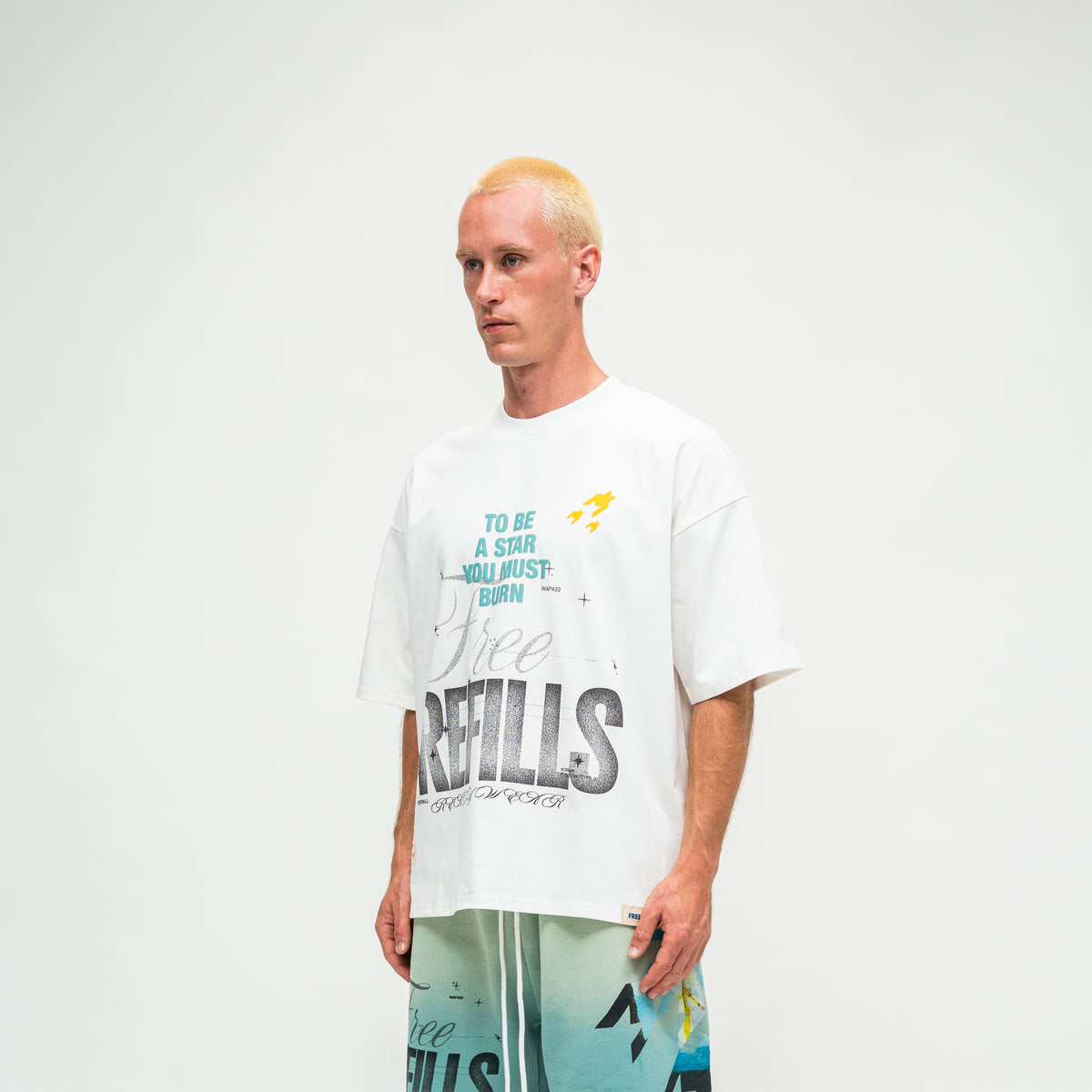 A model with bleached blonde hair wearing an oversized white graphic t-shirt and teal shorts, standing against a plain white studio background.