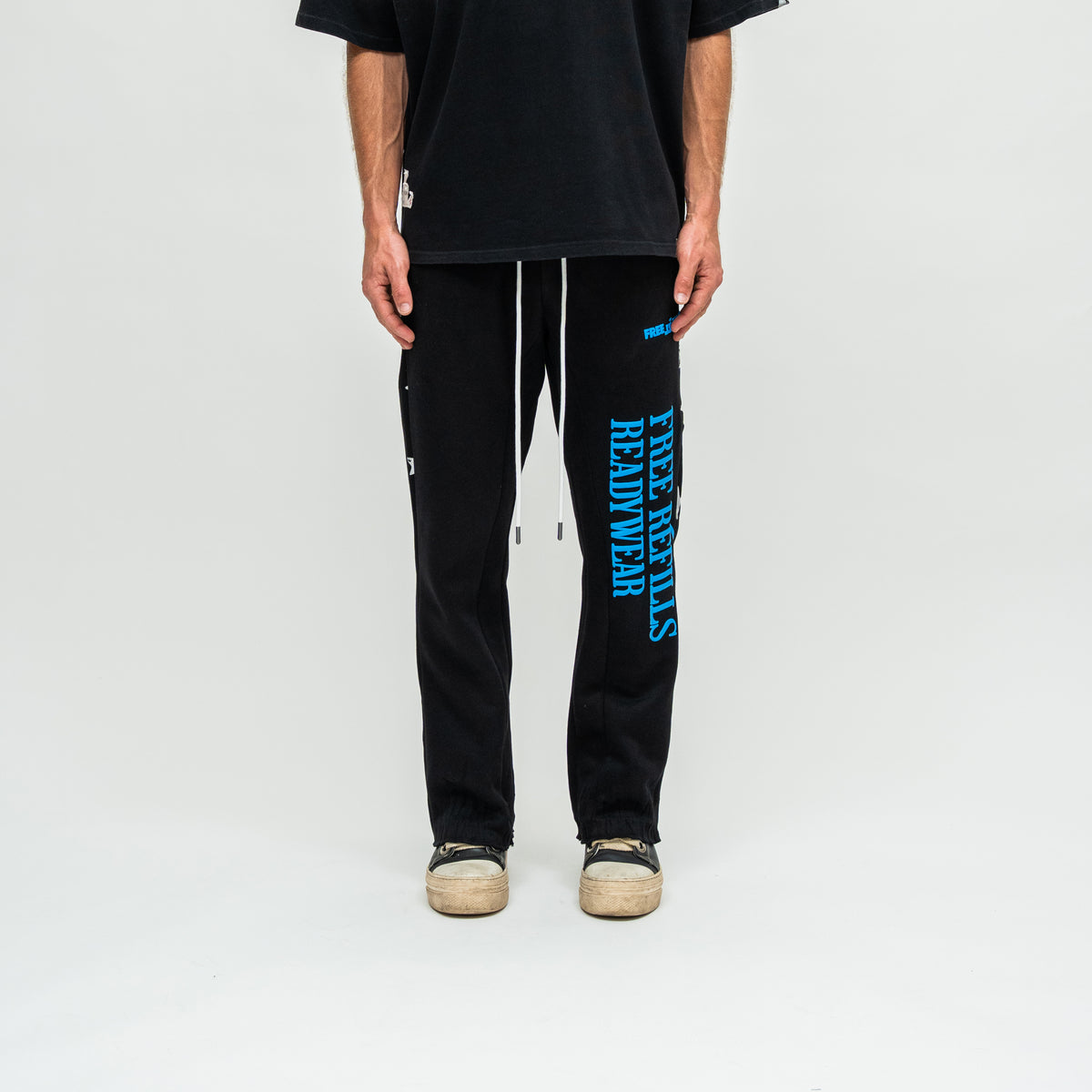 A person wearing black sweatpants with bright blue vertical text reading 'FREE REFILLS READYWEAR' on the left leg, a black t-shirt, and beige sneakers, photographed against a plain light gray studio background.