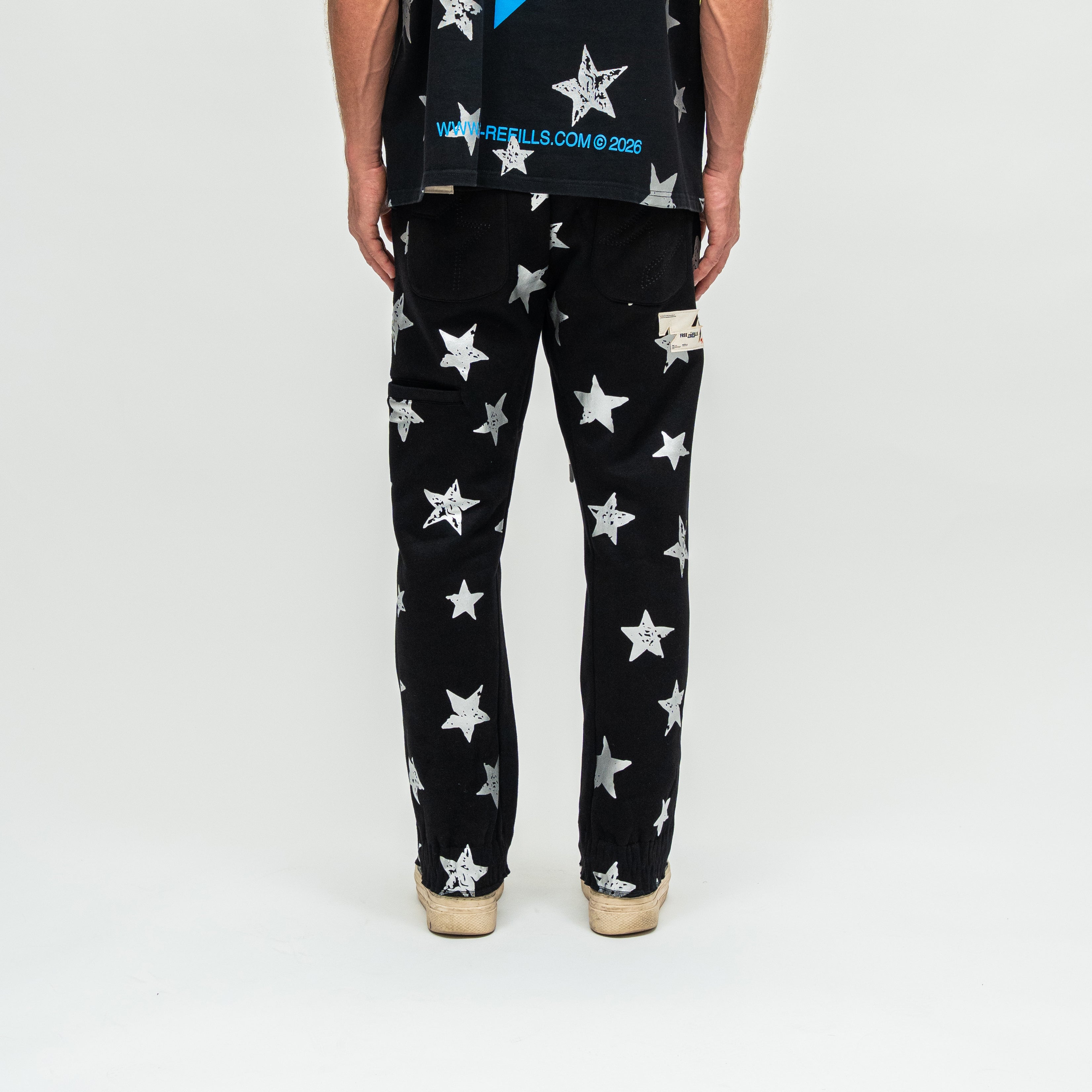 A person shown from the back wearing black pants with a white star print, a black graphic t-shirt, and beige sneakers, photographed against a plain white studio background.
