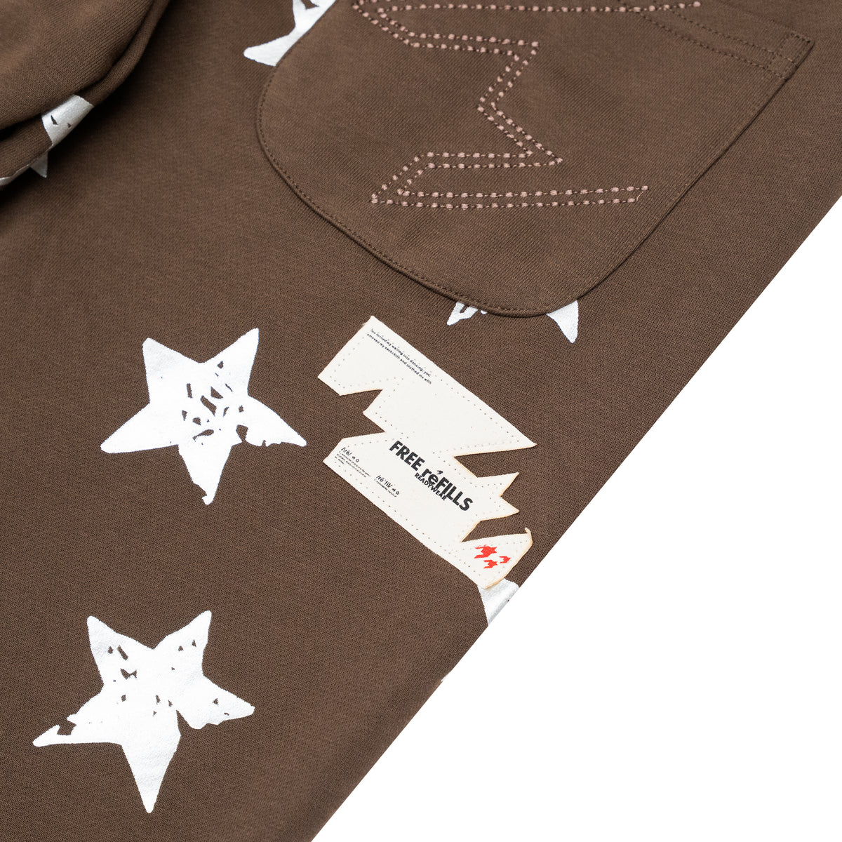 Close-up of a brown garment with white star graphics, a pocket with white stitched lightning-bolt detail, and a visible Free Refills brand tag.