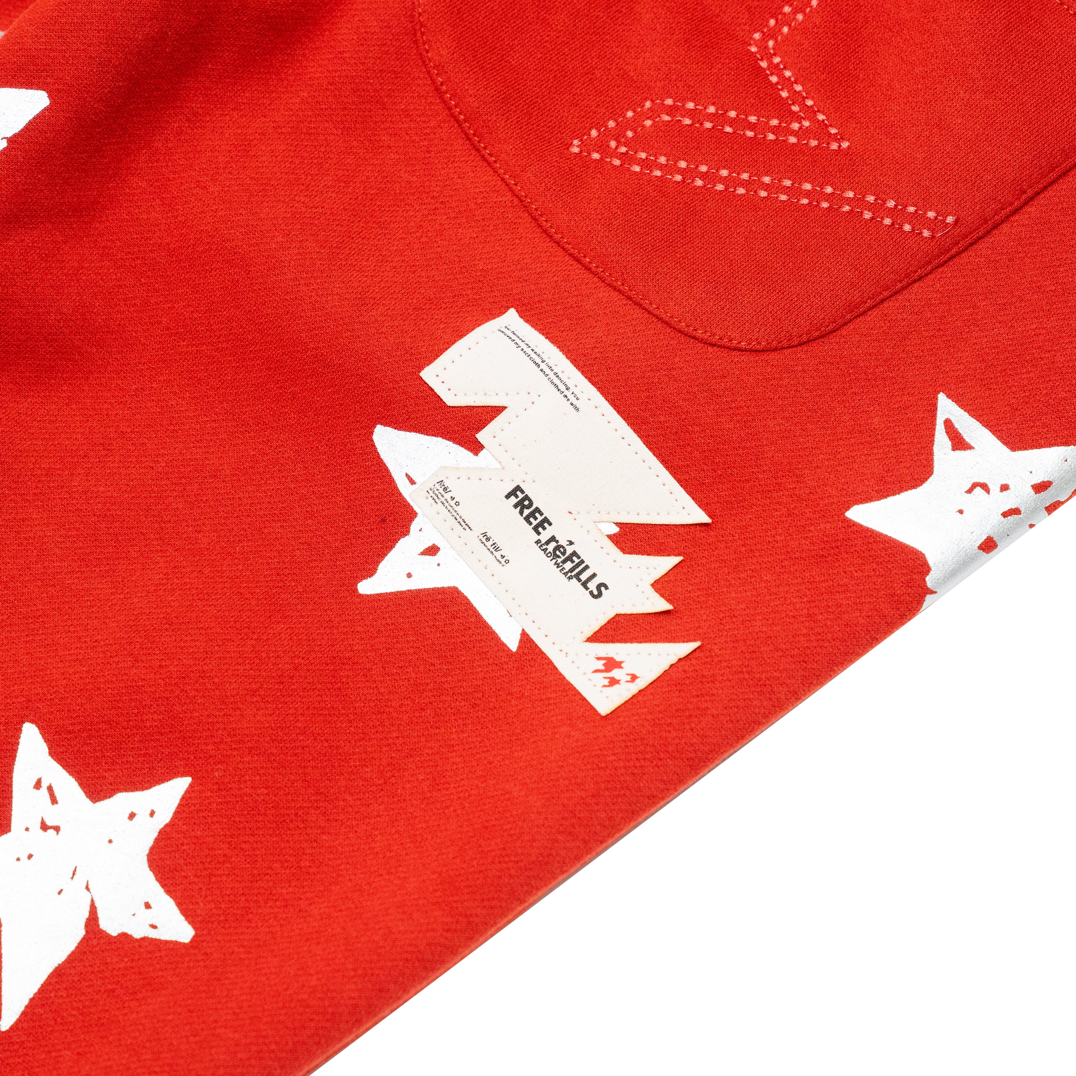 A close-up of a red garment, likely sweatpants or shorts, featuring a white graphic patch with the text 'FREE REFILLS' and white star motifs scattered across the fabric.