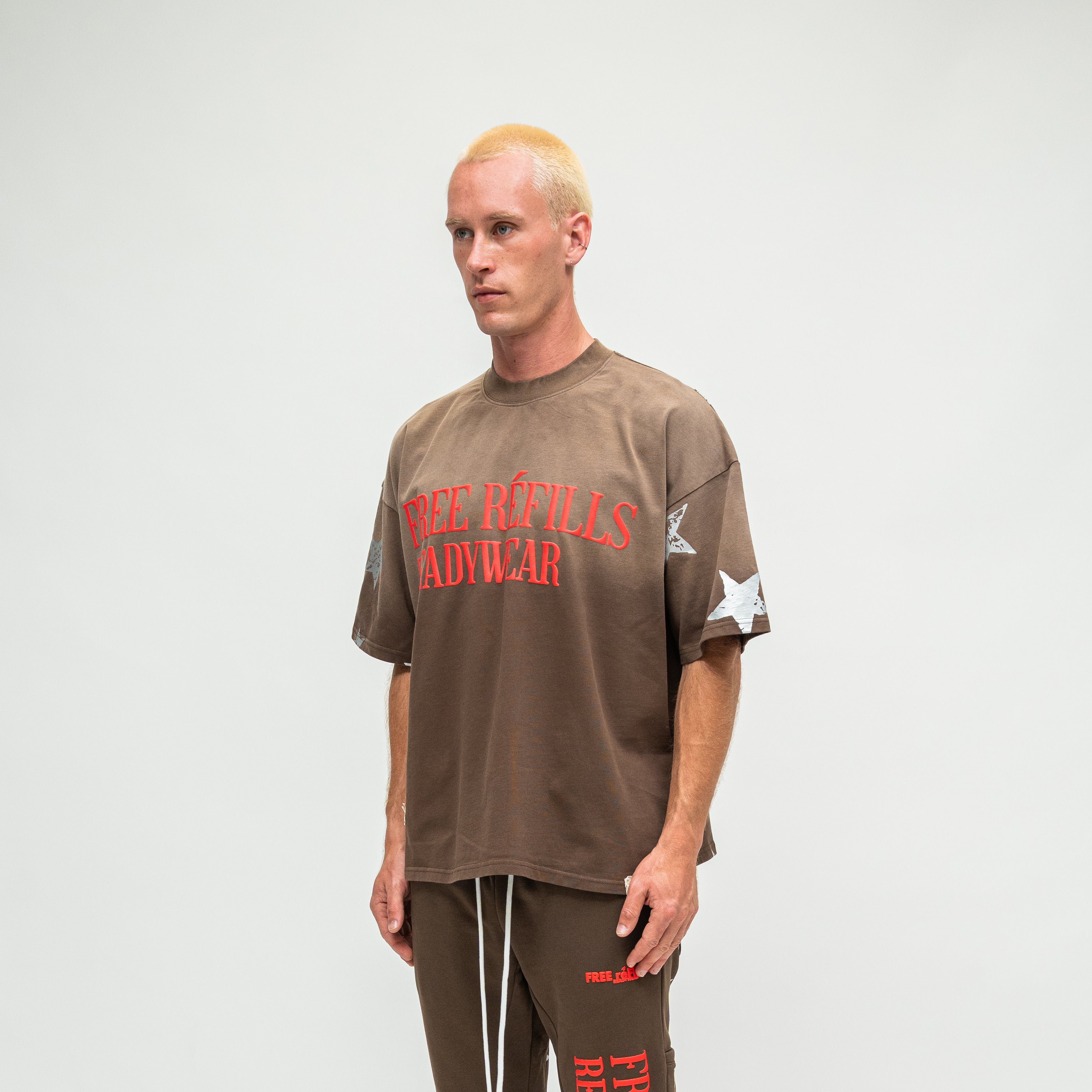 A man modeling a brown oversized t-shirt with 'FREE REFILLS READYWEAR' printed in red on the front, paired with matching brown sweatpants, against a plain light gray background.