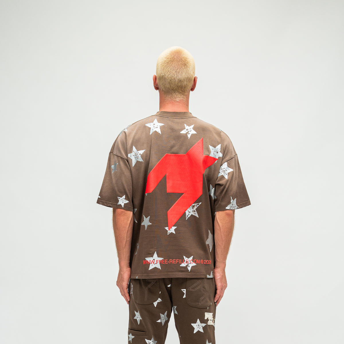 A man viewed from behind wearing a brown oversized t-shirt and matching pants, both covered in white star prints, with a large red graphic on the back of the shirt.