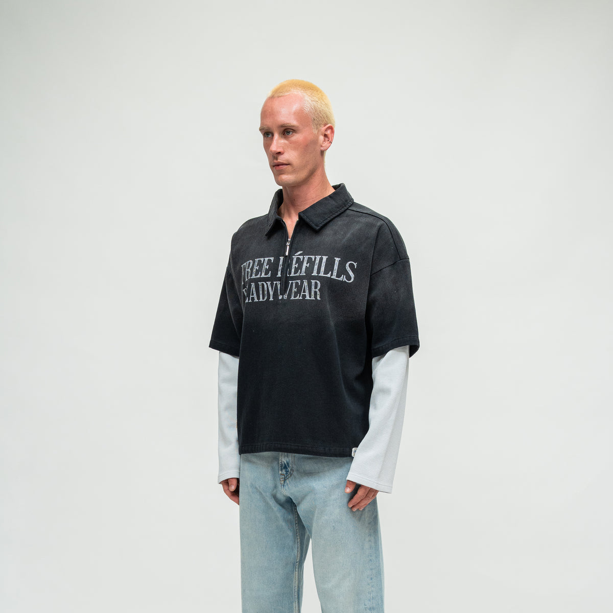 A model with bleached blonde hair stands against a light gray studio background wearing a black short-sleeve polo shirt with 'FREE REFILLS READYWEAR' printed in white across the chest, layered over a white long-sleeve shirt, and wearing light-wash blue jeans.