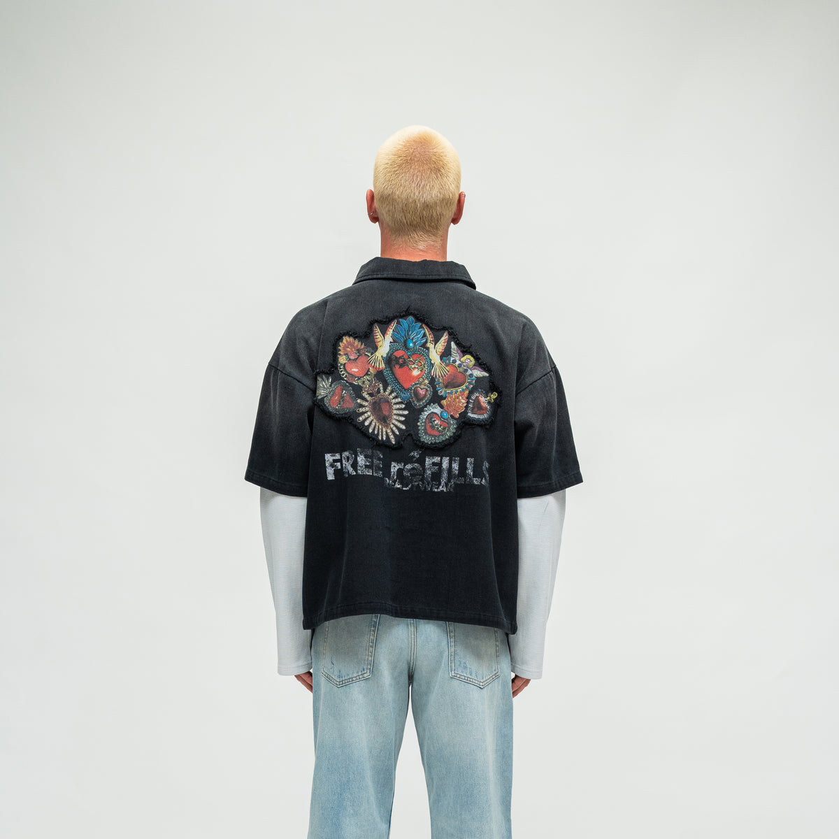 A model viewed from behind wearing a black short-sleeve button-up shirt with a large colorful graphic and 'FREE REFILLS' text on the back, layered over a white long-sleeve shirt and light blue jeans, against a light gray studio background.