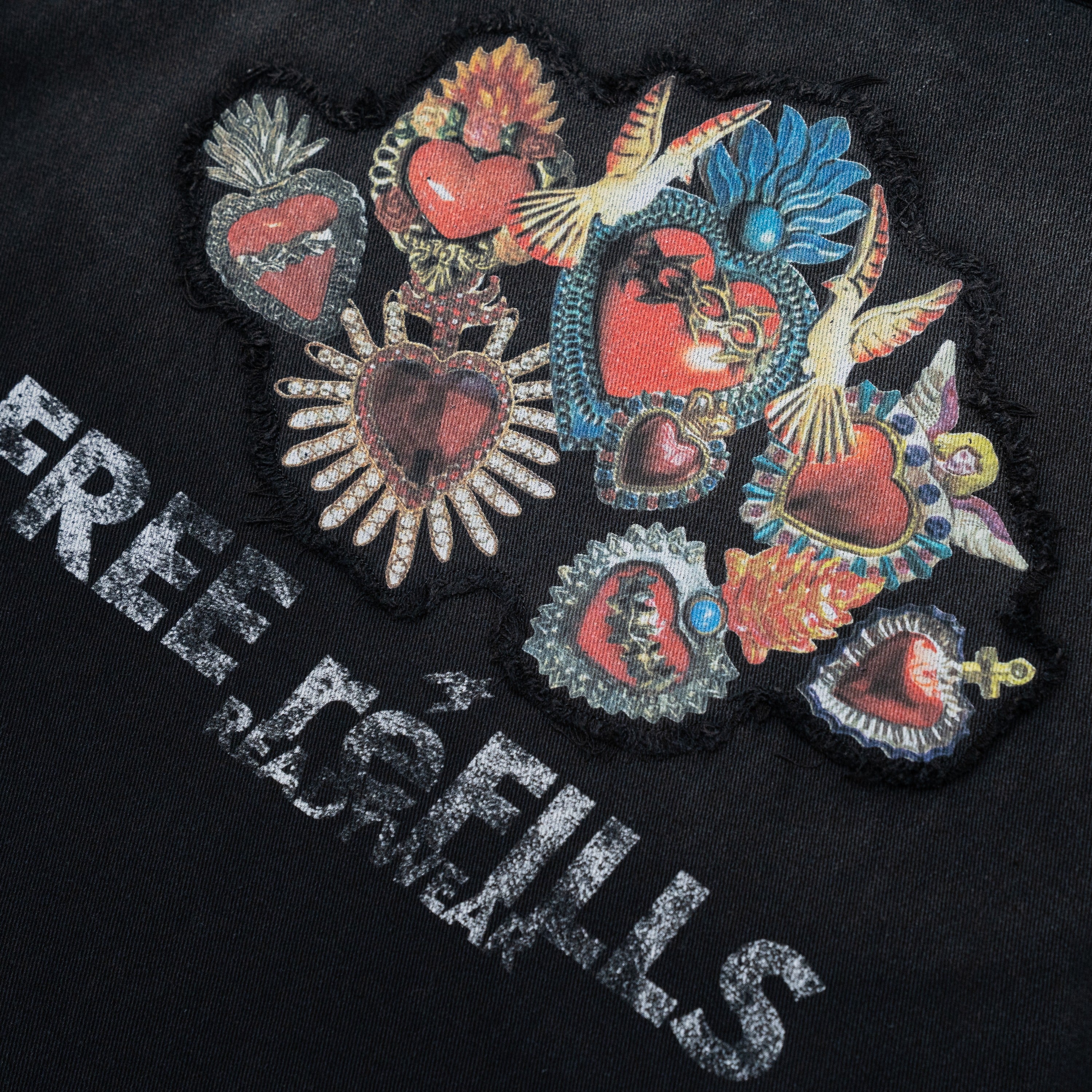 Close-up of a black garment featuring a large multicolor patchwork design of ornate red hearts, birds, and floral motifs, with distressed white text reading 'FREE REFILLS' across the lower left.