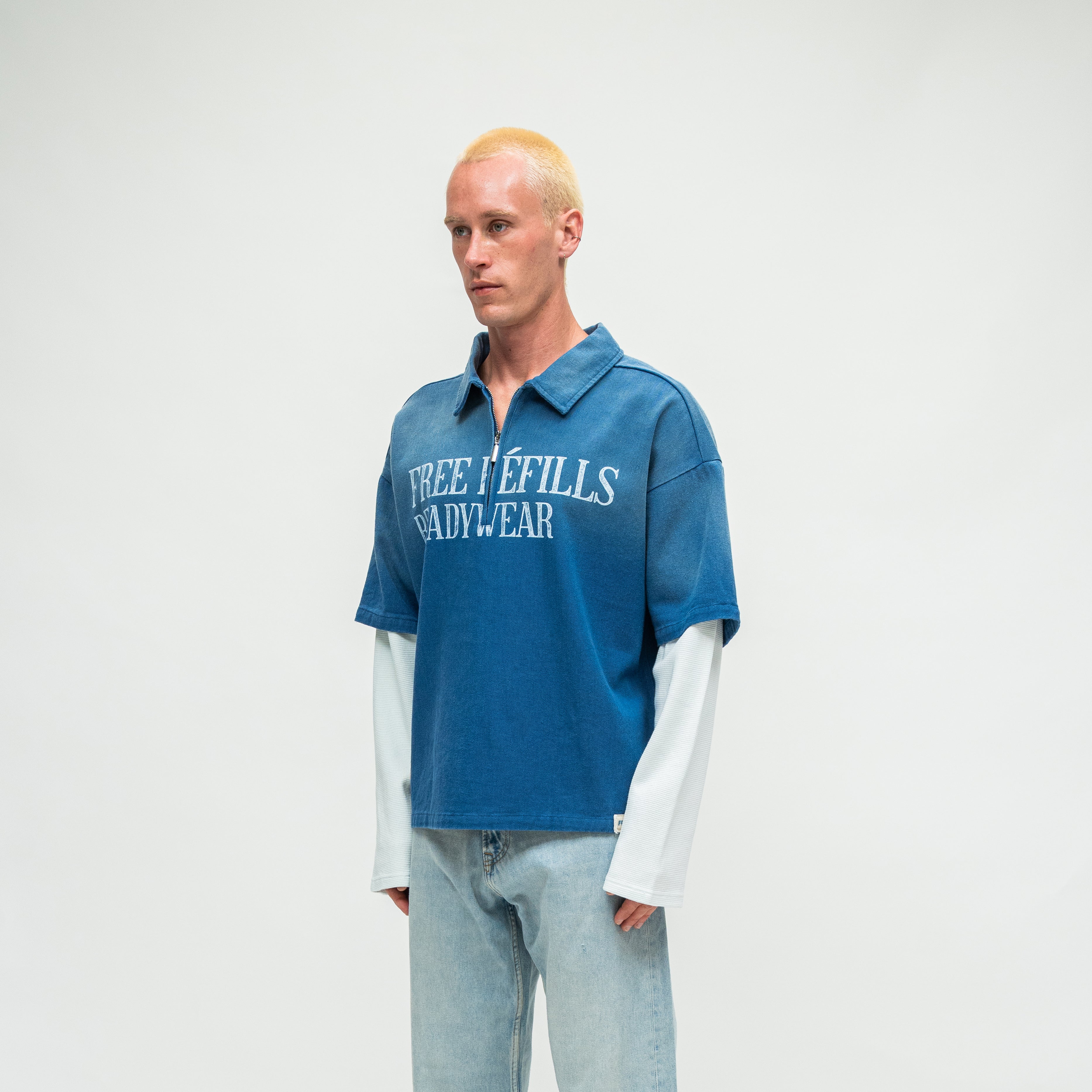 A model with bleached blonde hair stands against a light gray studio background wearing a blue short-sleeve polo shirt with 'FREE REFILLS READYWEAR' printed in white across the chest, layered over a white long-sleeve shirt and light-wash jeans.