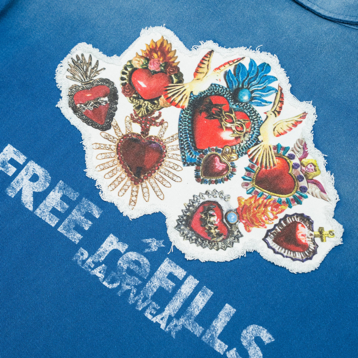 Close-up of a blue graphic T-shirt featuring a large white patch with multiple red hearts, gold and blue decorative details, and distressed white text reading 'FREE REFILLS' across the chest.