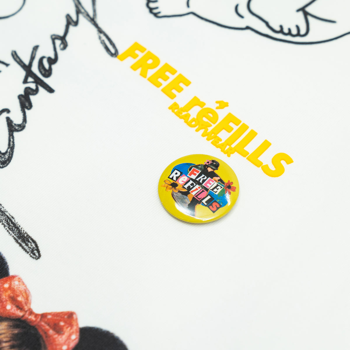 The Powder Room Tee – Cream – Close-up of yellow FREE reFILLS lettering and a round graphic pin
