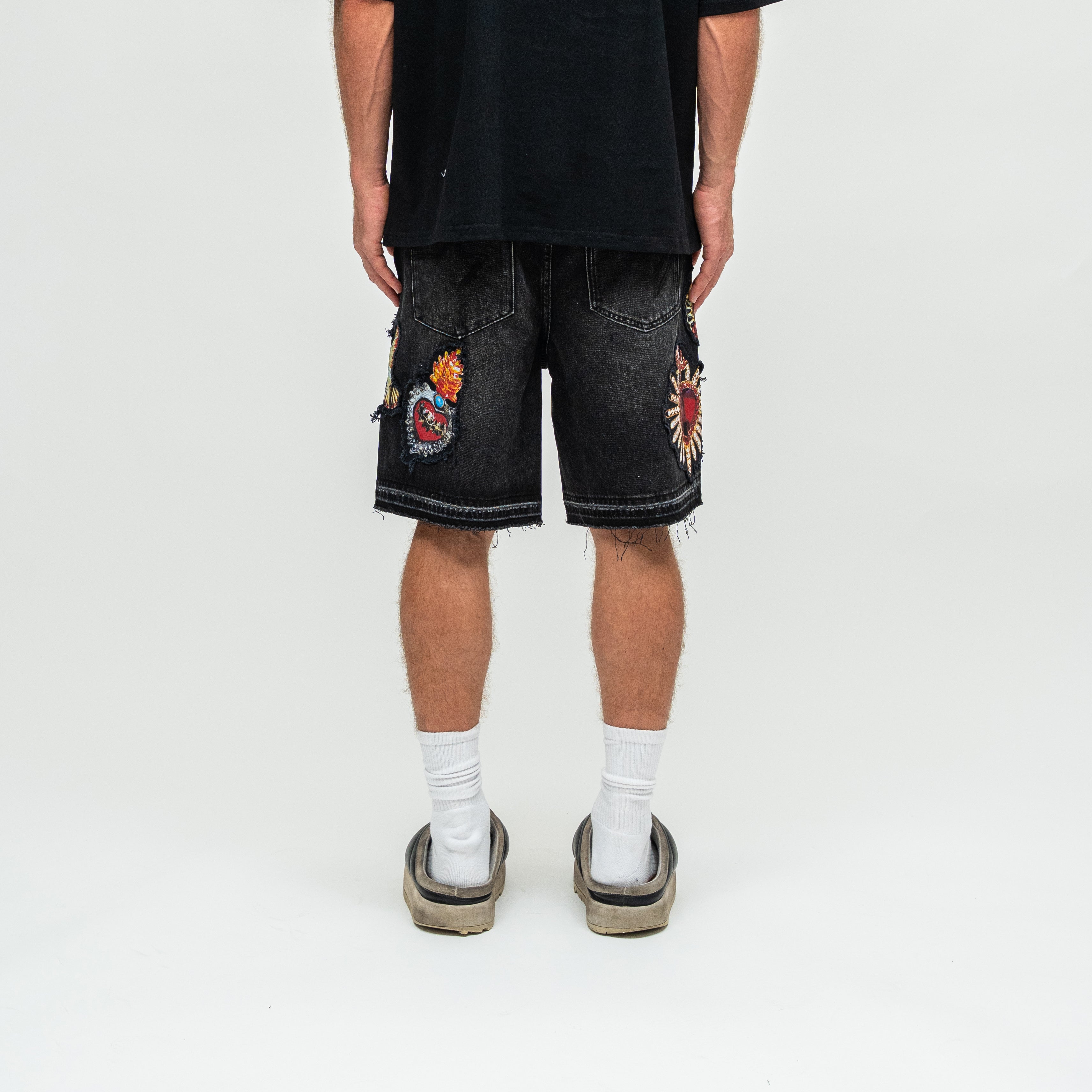 A person shown from the back wearing black denim shorts with colorful embroidered patches, a black t-shirt, white socks, and black-and-white platform sandals on a white studio background.