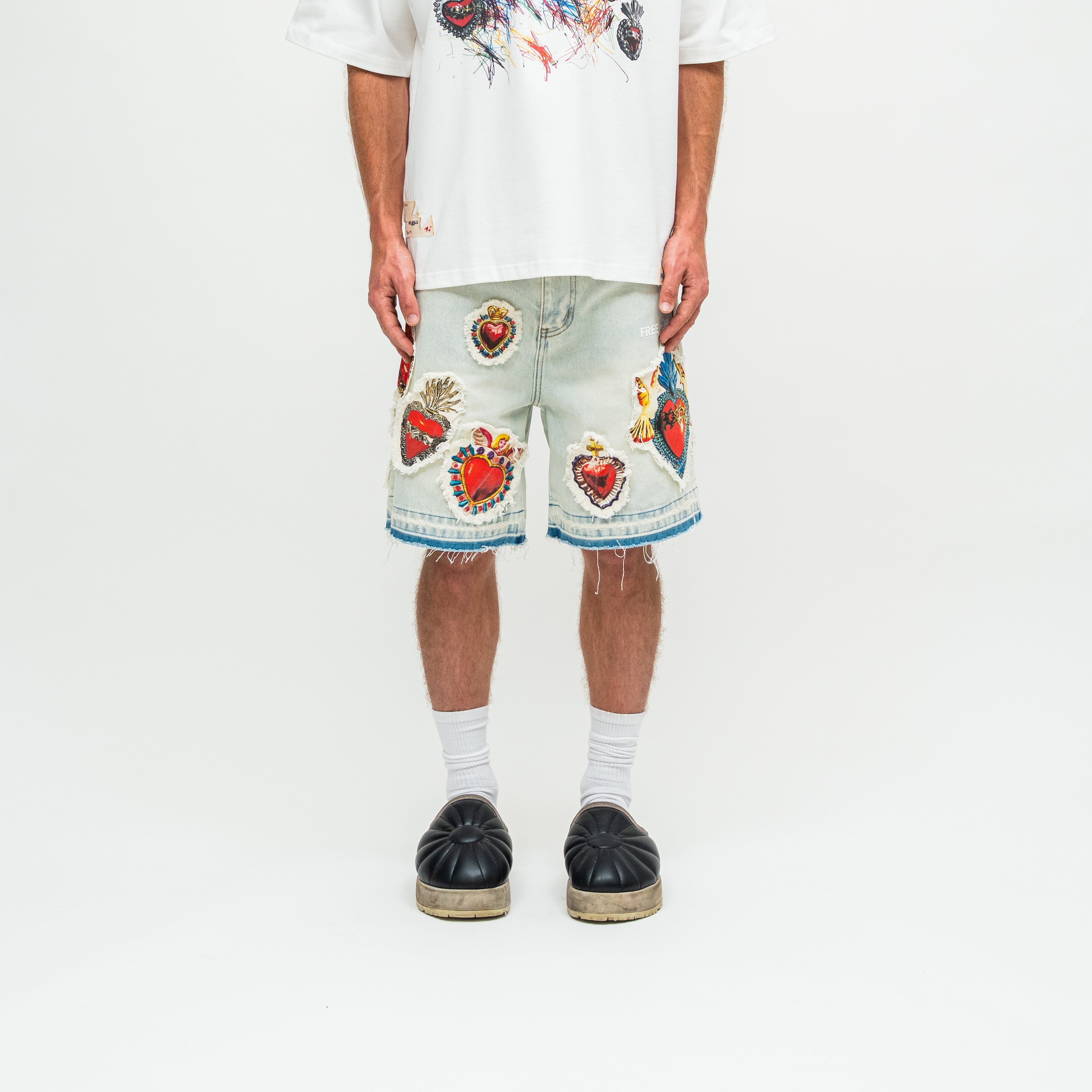 A model is shown from the waist down wearing light-wash denim shorts heavily decorated with colorful, detailed religious heart patches, paired with a white graphic t-shirt, white socks, and black espadrille platform shoes against a plain white studio background.