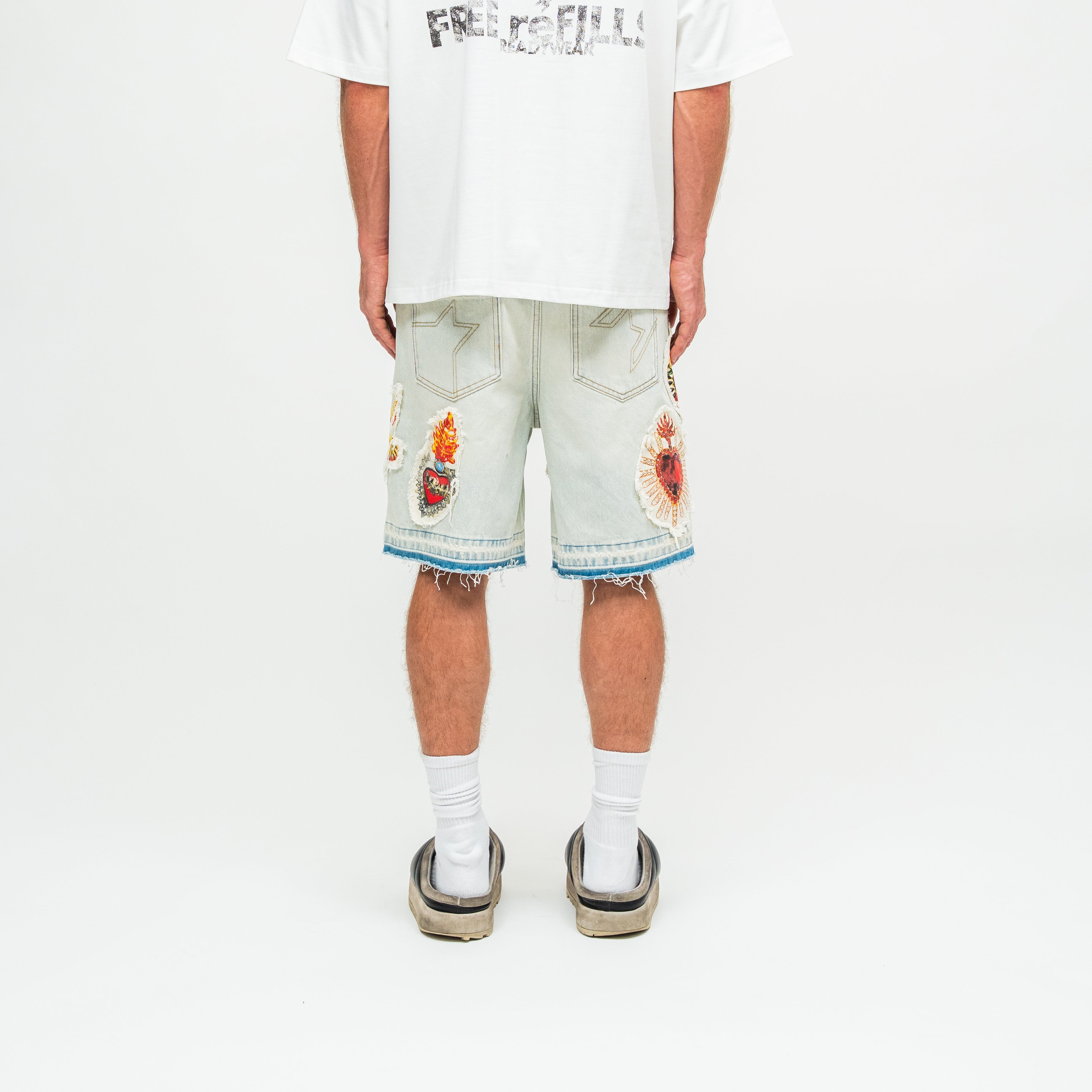 A model shown from the back wearing light-wash denim shorts with colorful embroidered patches on the back pockets and frayed hems, styled with a white graphic t-shirt, white socks, and black slide sandals against a plain white studio background.