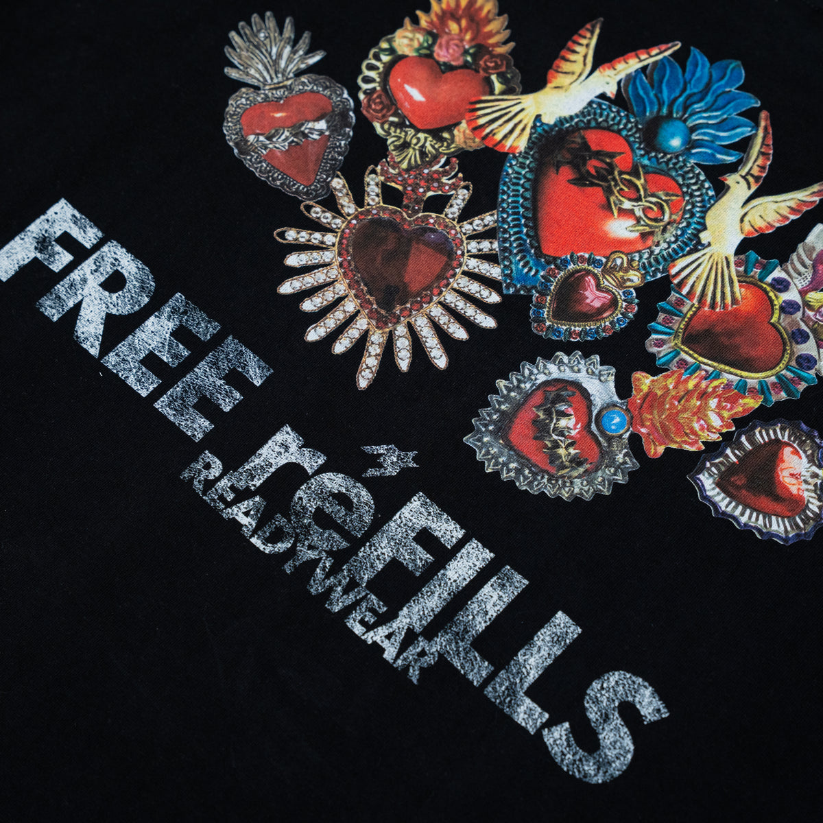 A close-up of a black graphic T-shirt featuring the text 'FREE REFILLS' in distressed silver lettering, alongside a cluster of colorful religious-style heart medallions and ornate emblems.