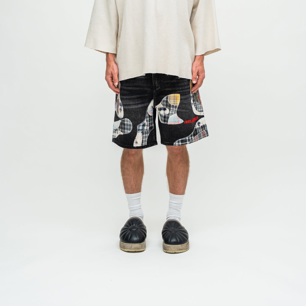 A model is shown from the waist down wearing black denim shorts with a white plaid and beige abstract patchwork print, styled with white socks and black platform slip-on shoes against a plain white studio background.