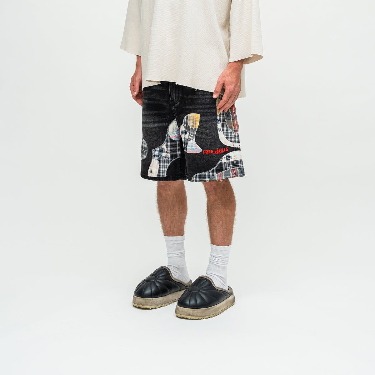 A person wearing black denim shorts with a patchwork design of plaid patterns and red text, styled with white socks and black espadrille mule shoes against a plain white studio background.