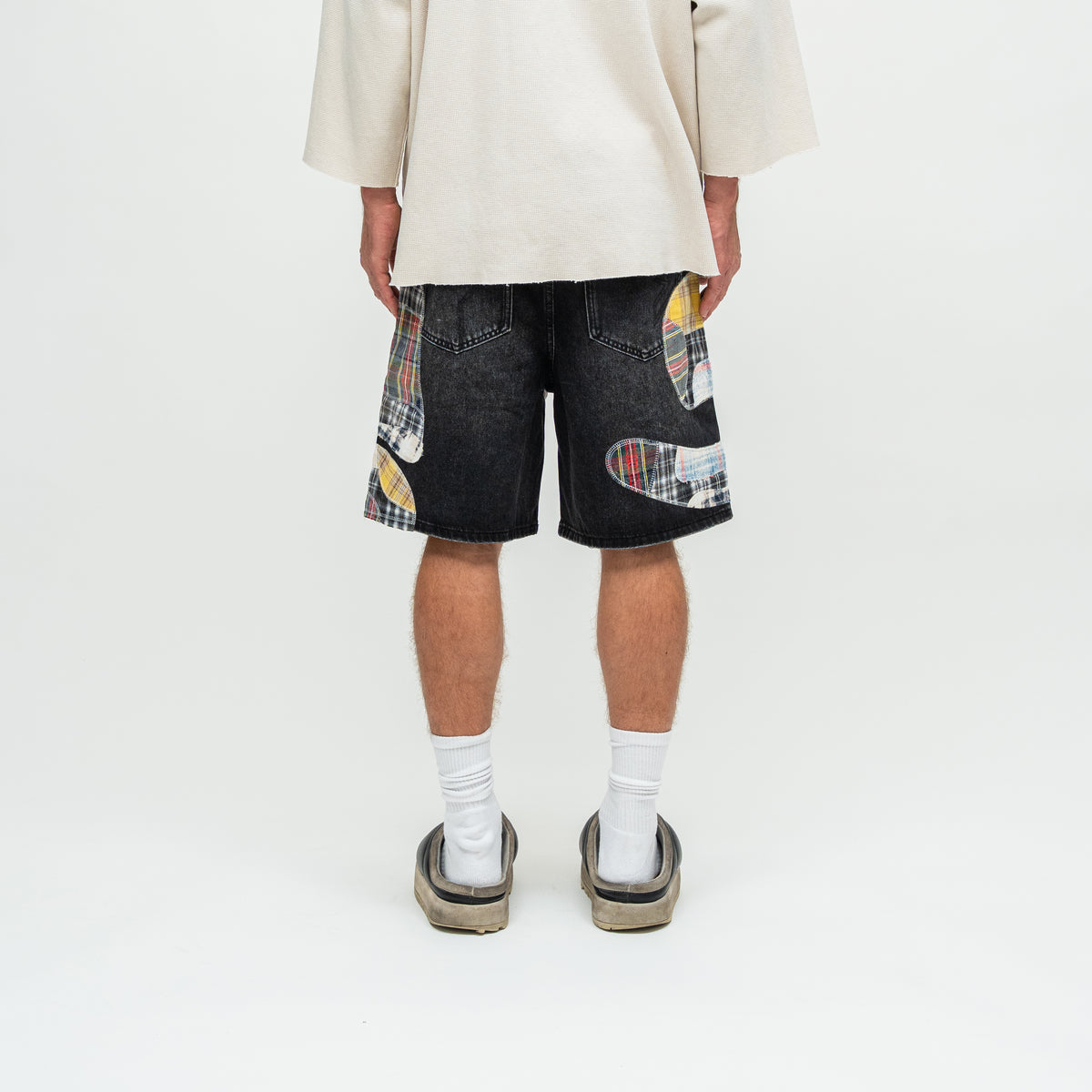 A person shown from the back wearing dark denim shorts with colorful patchwork skateboard graphics, a cream-colored shirt, white socks, and slip-on shoes against a plain white studio background.