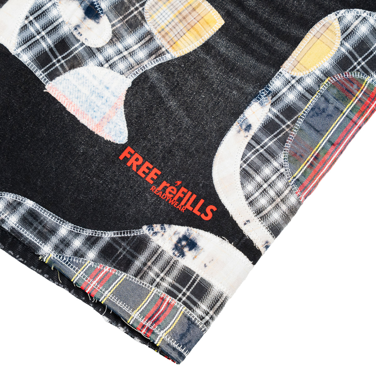 A close-up of a denim garment corner showing a black denim base with a patchwork applique design of plaid and tie-dye fabrics in white, yellow, red, and blue, featuring the red text 'FREE REFILLS READYWEAR'.