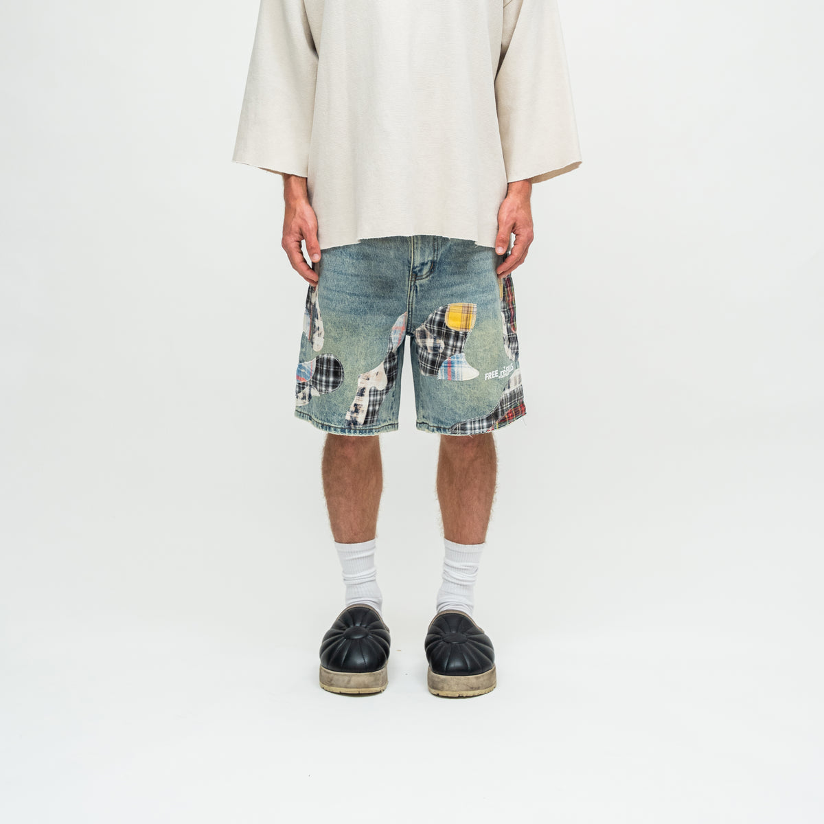 A person shown from the chest down wearing distressed denim shorts with a patchwork design, a cream-colored oversized t-shirt, white socks, and black platform sandals against a plain white studio background.