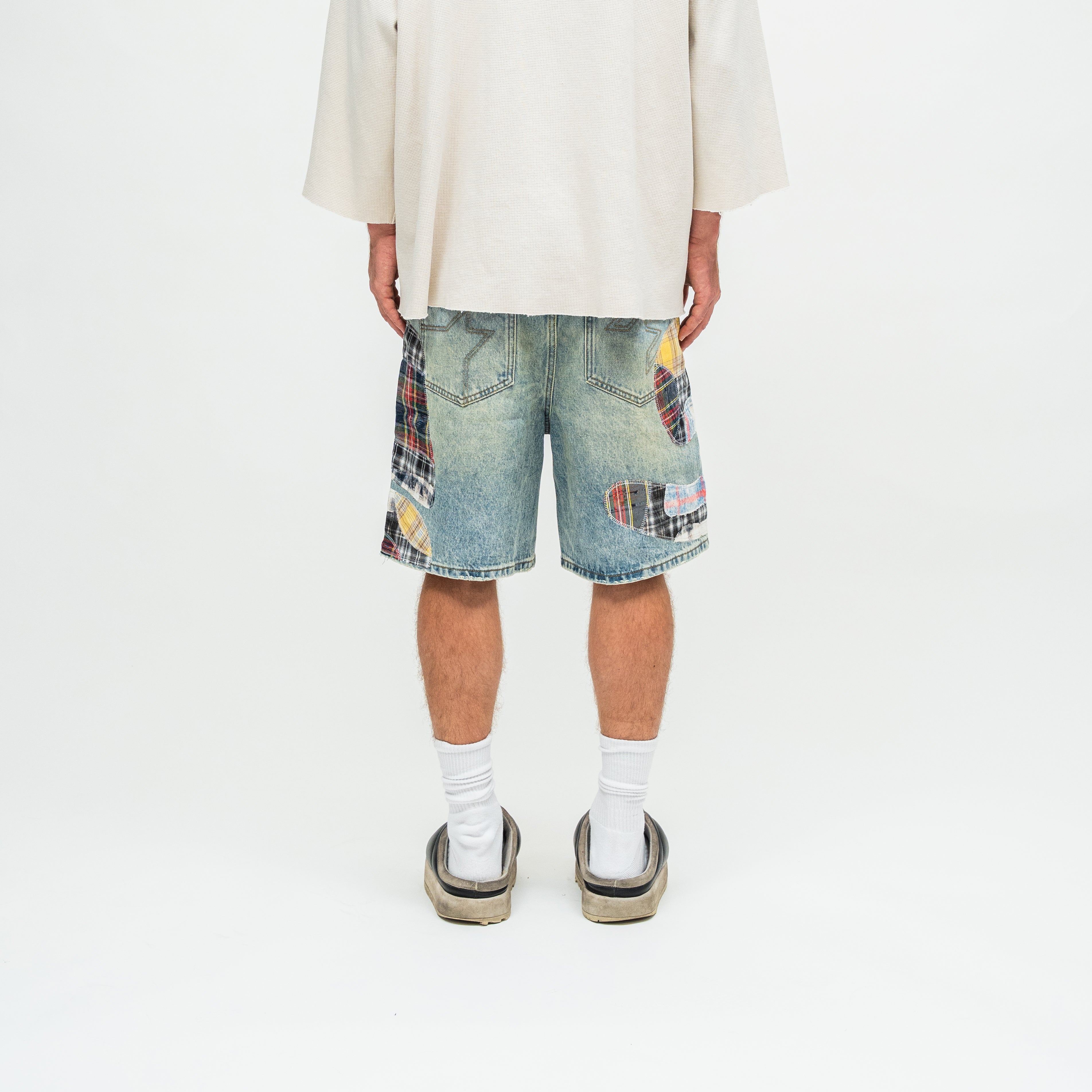 A person shown from the back wearing light-wash denim shorts with colorful patchwork plaid details on the back pockets, a cream-colored oversized shirt, white socks, and platform sandals, against a plain white studio background.