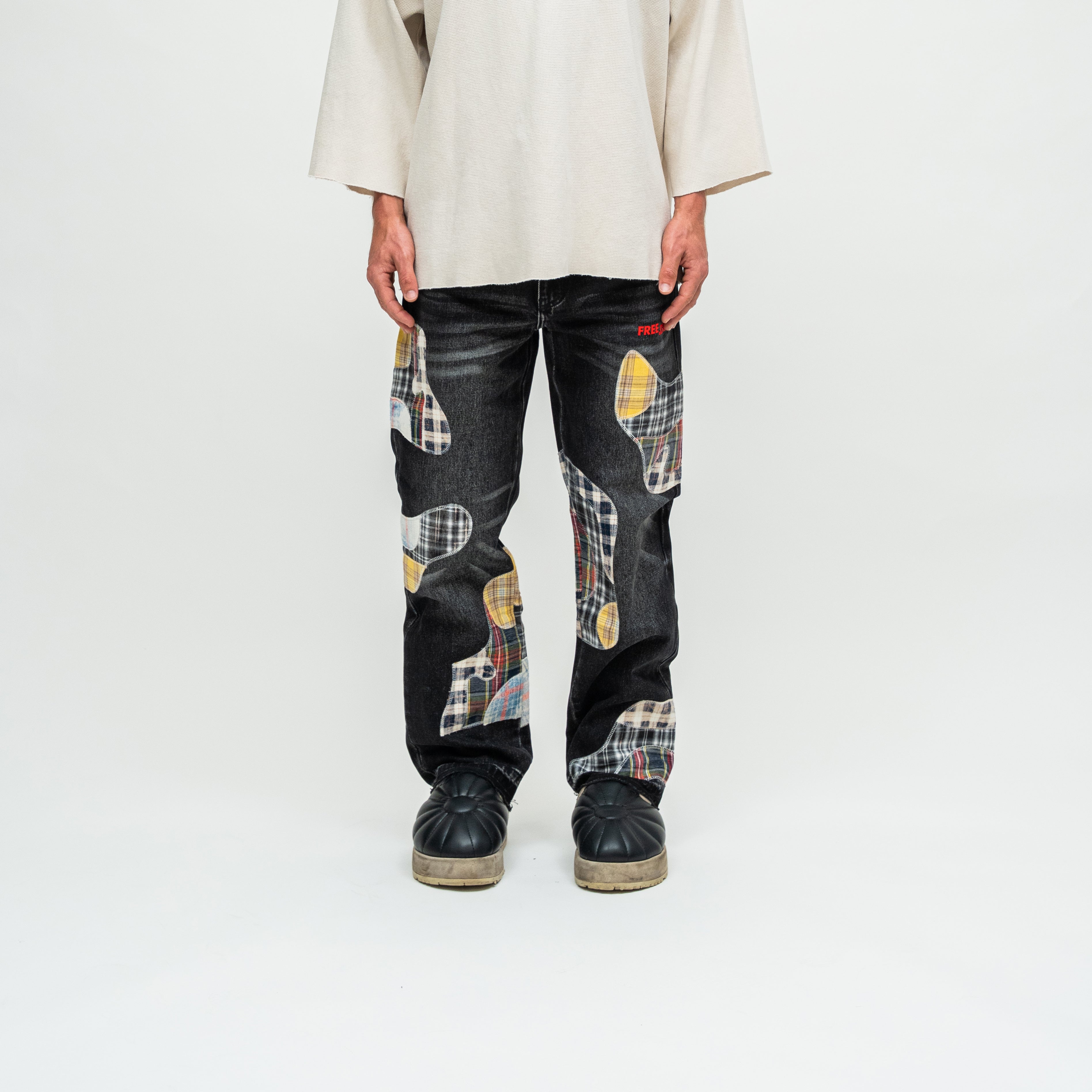 A person standing against a light gray studio background wearing dark patchwork jeans with plaid and abstract color-block patches, a loose cream top, and black shoes.