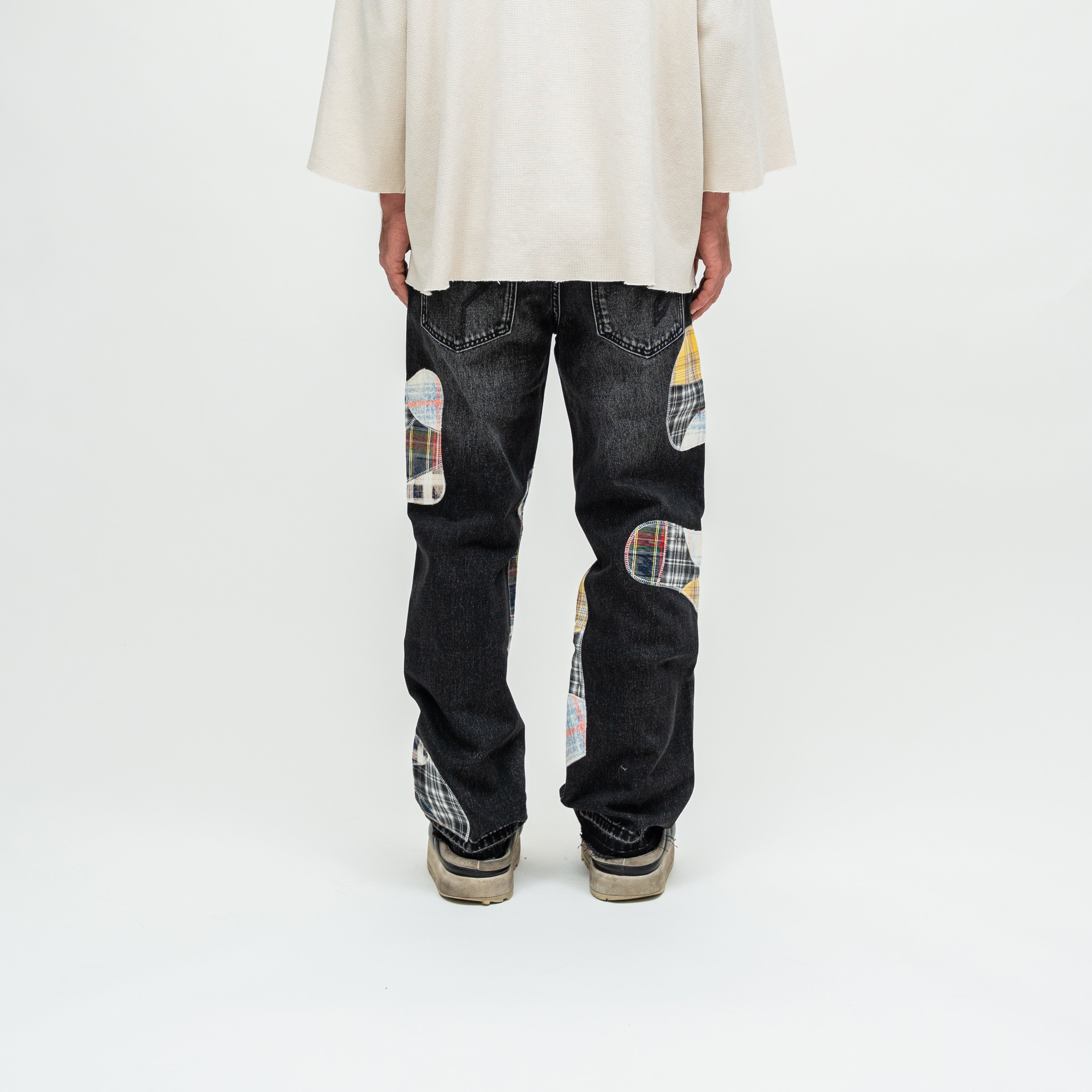A person shown from the back wearing dark patchwork jeans with plaid knee patches and a cream oversized top, photographed against a clean white studio background.