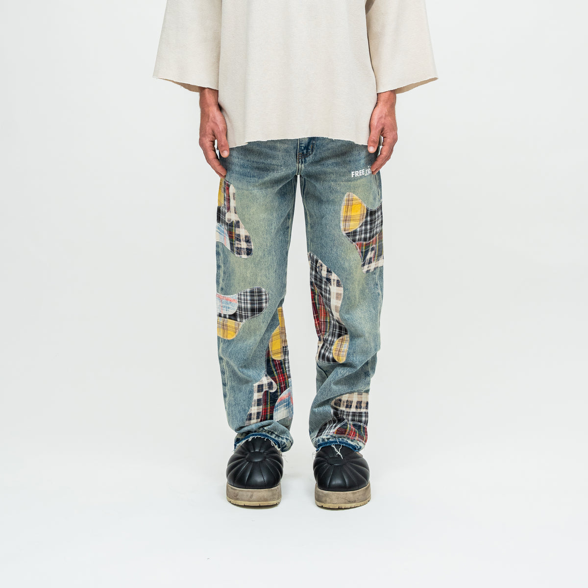 A person wearing loose-fit blue denim jeans with large multicolor patchwork plaid inserts, paired with a cream shirt and black shoes, photographed against a plain white studio background.