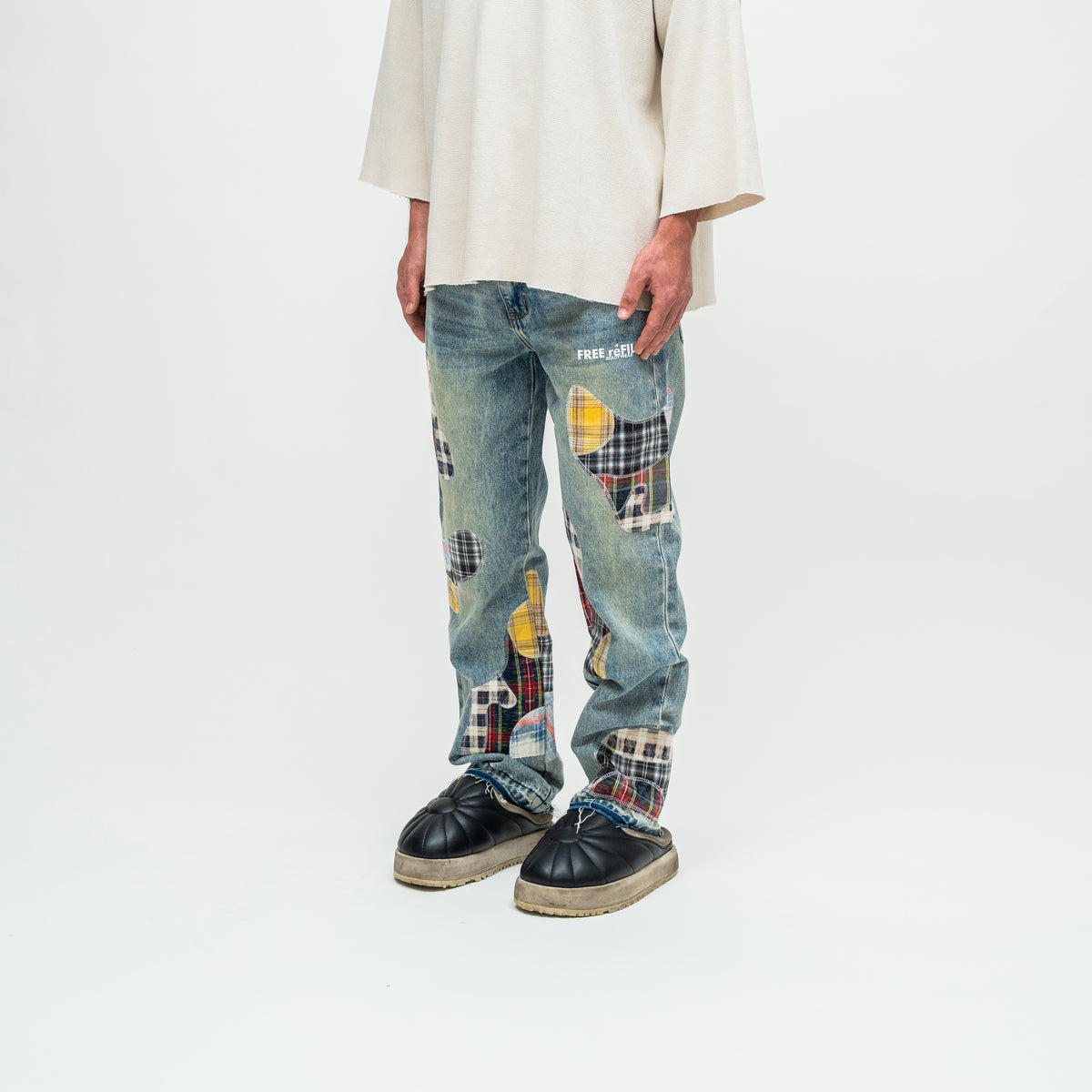 A person wearing light blue patchwork denim jeans with yellow, red, and black plaid details, a cream-colored oversized top, and black leather shoes, photographed against a plain white studio background.