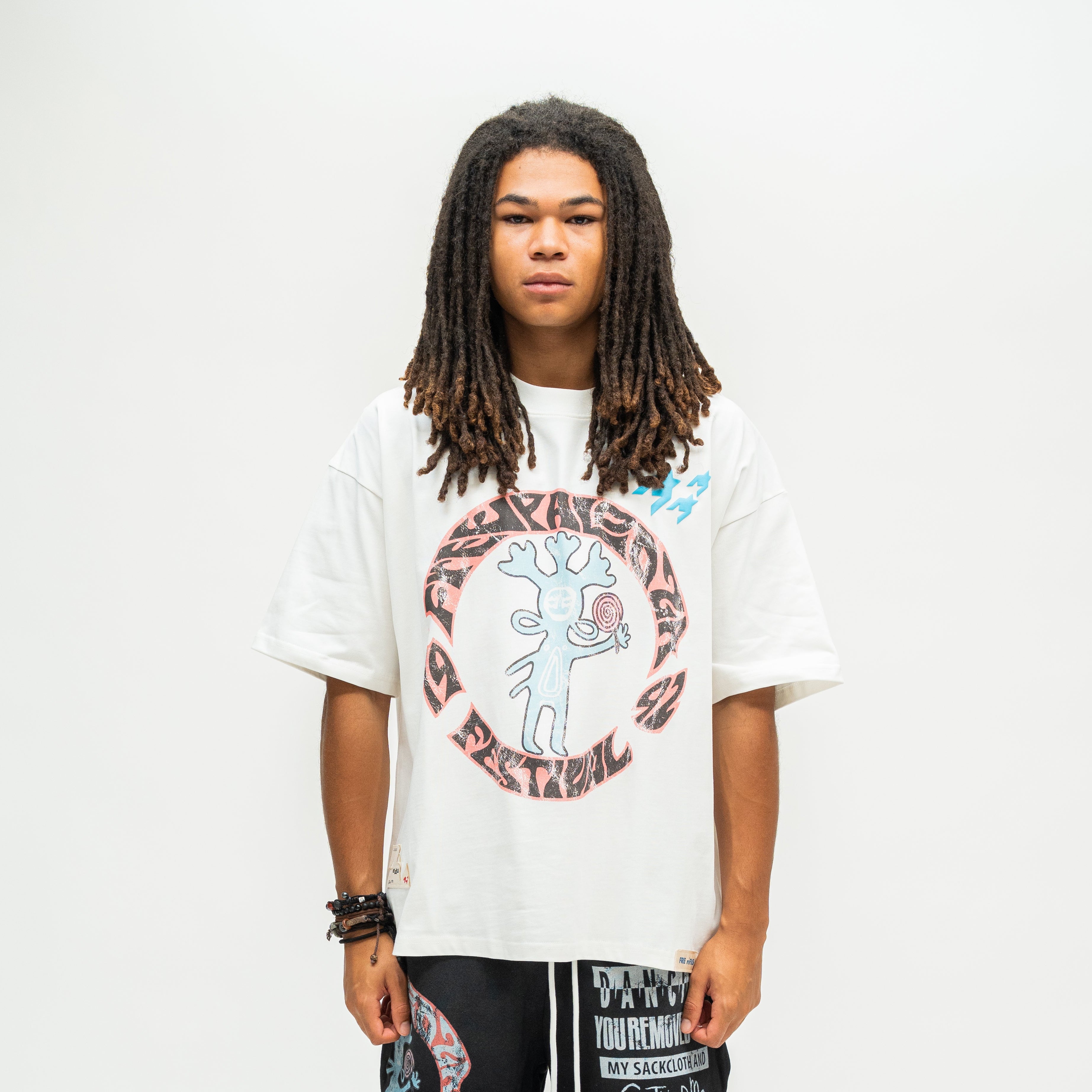 A person with long dreadlocks stands against a plain light background wearing an oversized white graphic T-shirt with a circular red, black, and blue print, paired with black patterned pants.