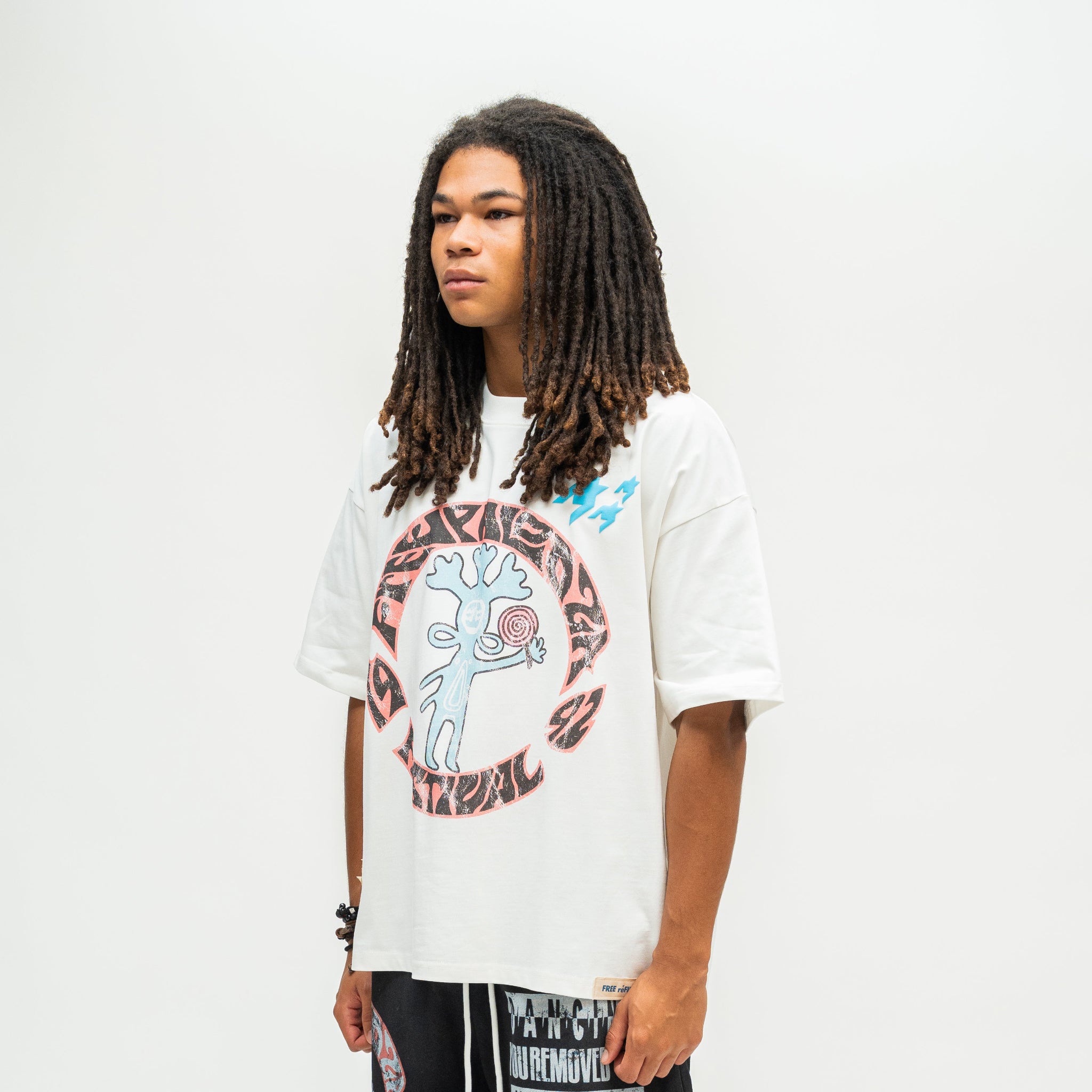 A person with long dreadlocks stands against a plain light gray background wearing a white graphic T-shirt and black pants with white text graphics.