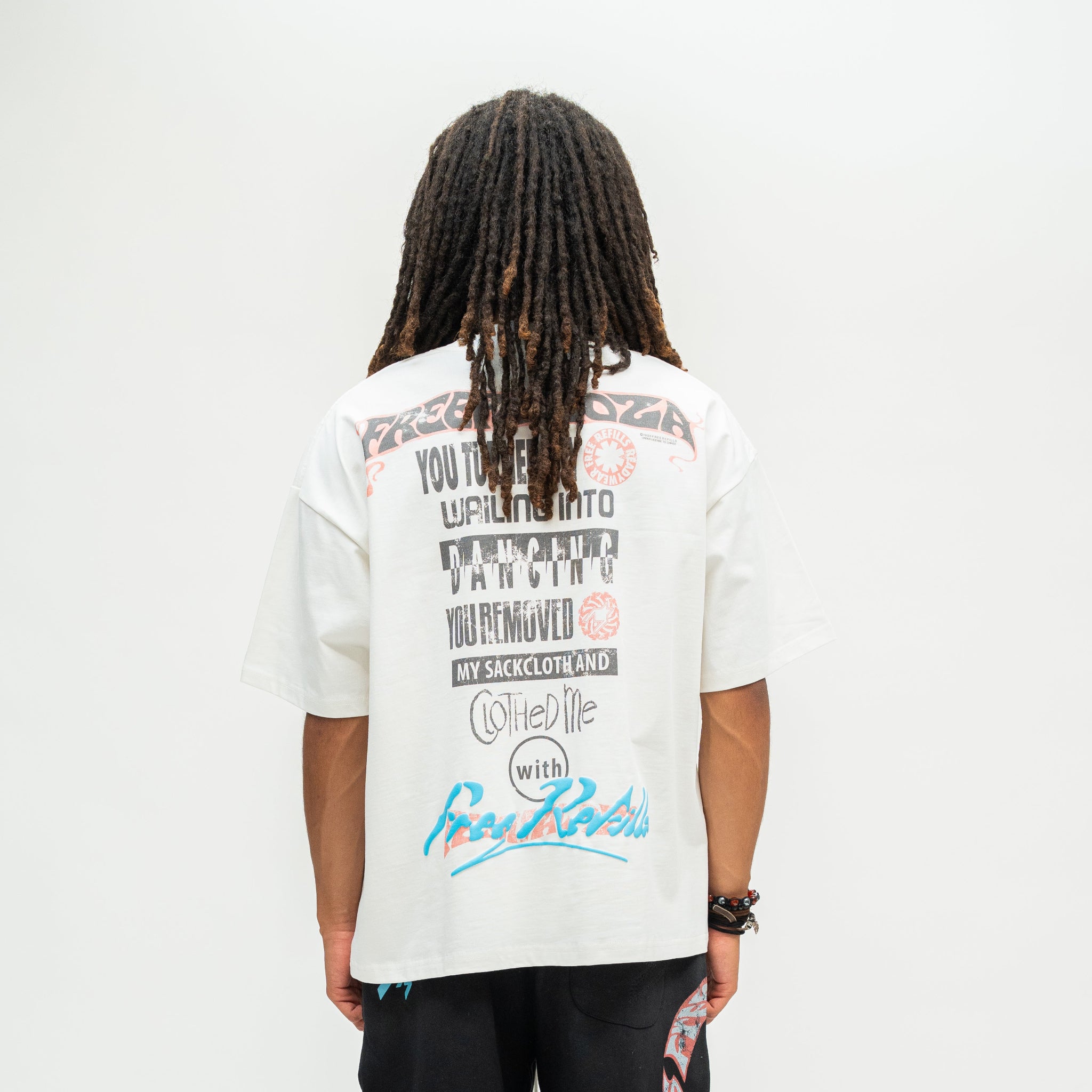 A person viewed from behind wearing a white graphic T-shirt with black, pink, and blue text and artwork, paired with black pants, against a plain light gray studio background.