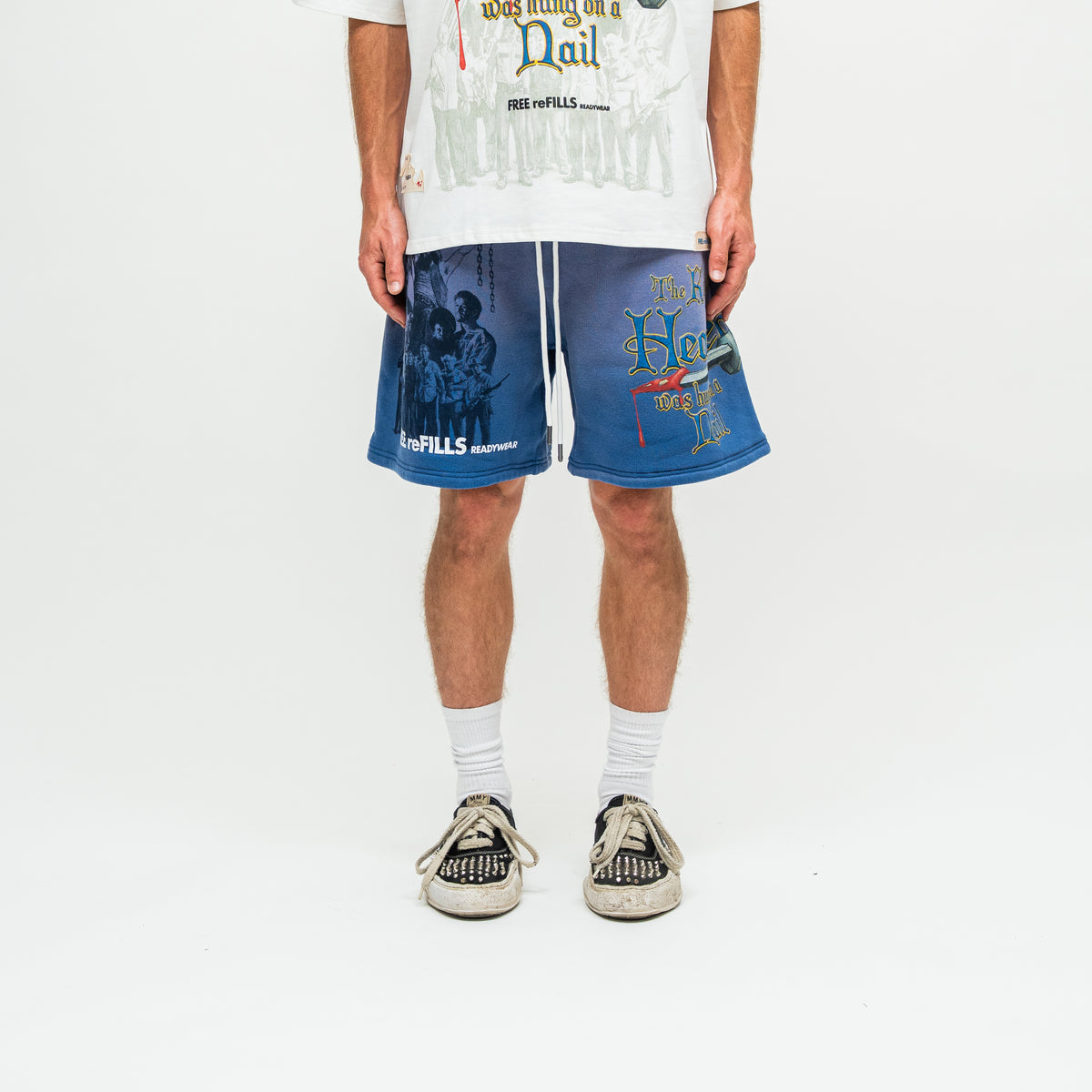 A person wearing blue graphic sweat shorts with white drawstrings, a white graphic t-shirt, white socks, and black-and-white sneakers, standing against a plain white studio background.