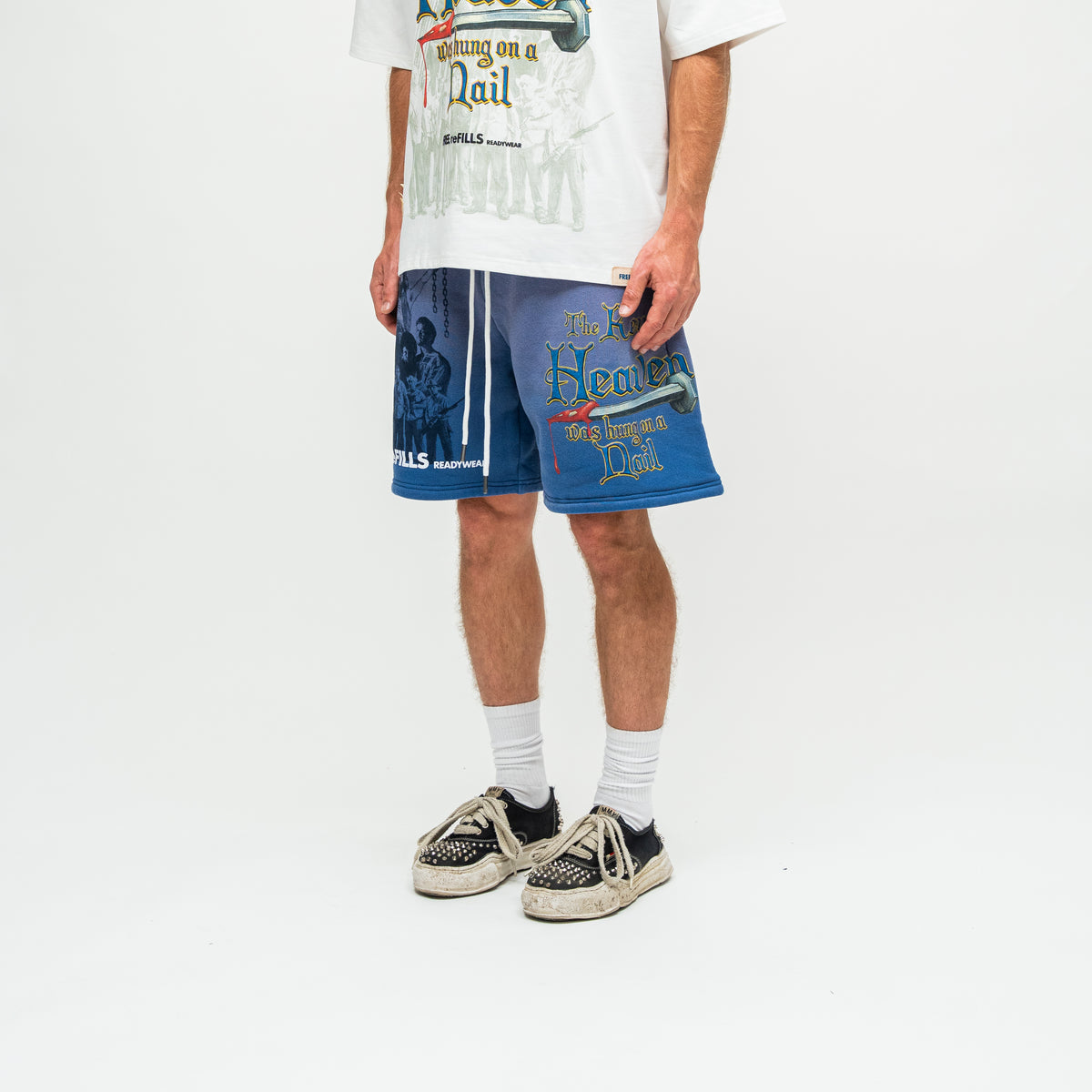 A person wearing blue graphic drawstring shorts, a white graphic t-shirt, white socks, and black-and-white sneakers, photographed against a clean white studio background.