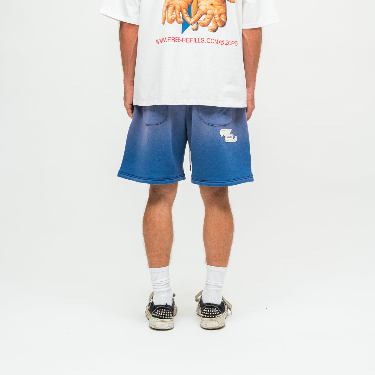 A person shown from the back wearing blue gradient sweat shorts with a small white logo, a white graphic t-shirt, white socks, and black sneakers on a white studio background.