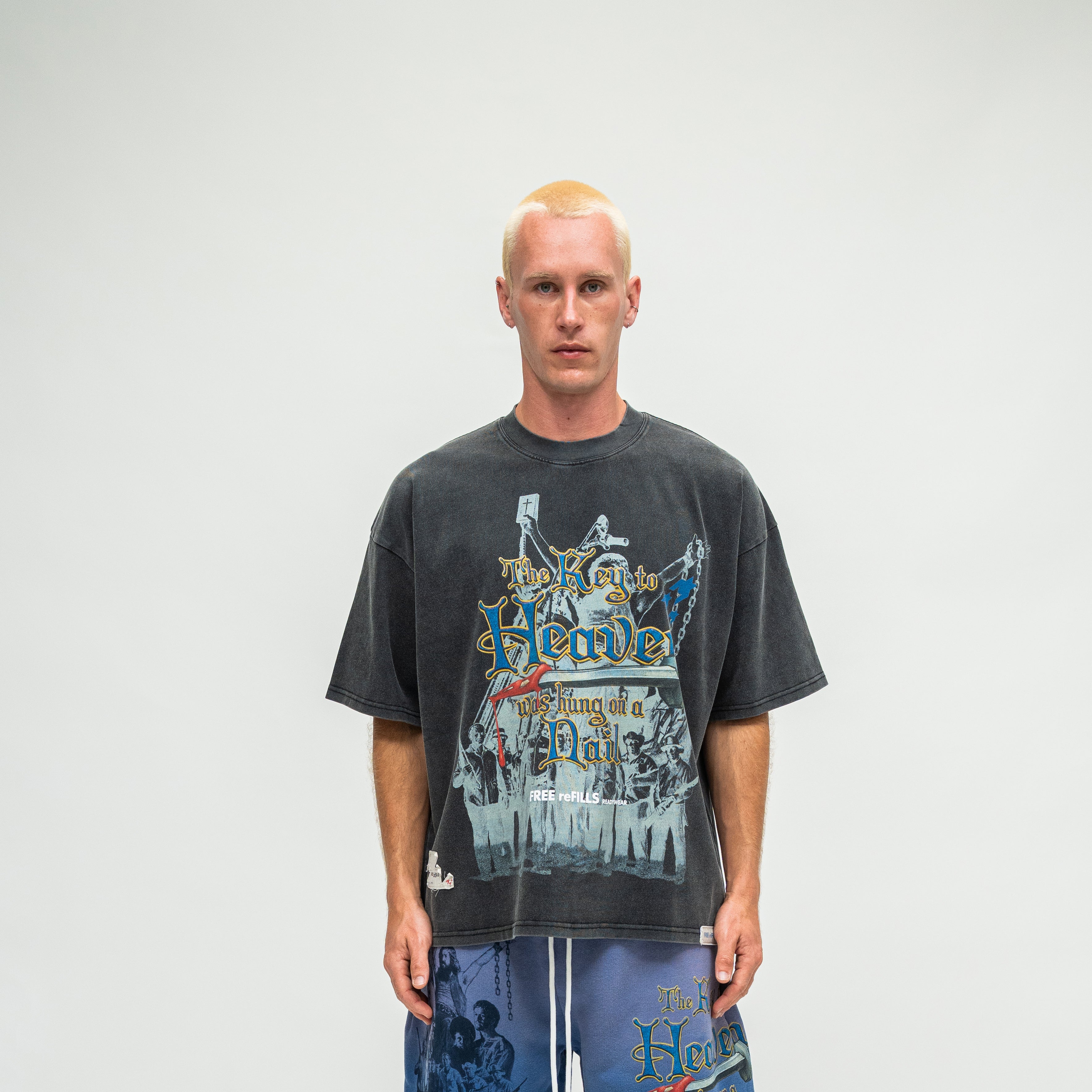 A man wearing a faded black graphic t-shirt with a vintage-style illustration and text, paired with blue shorts, standing against a plain light gray background.
