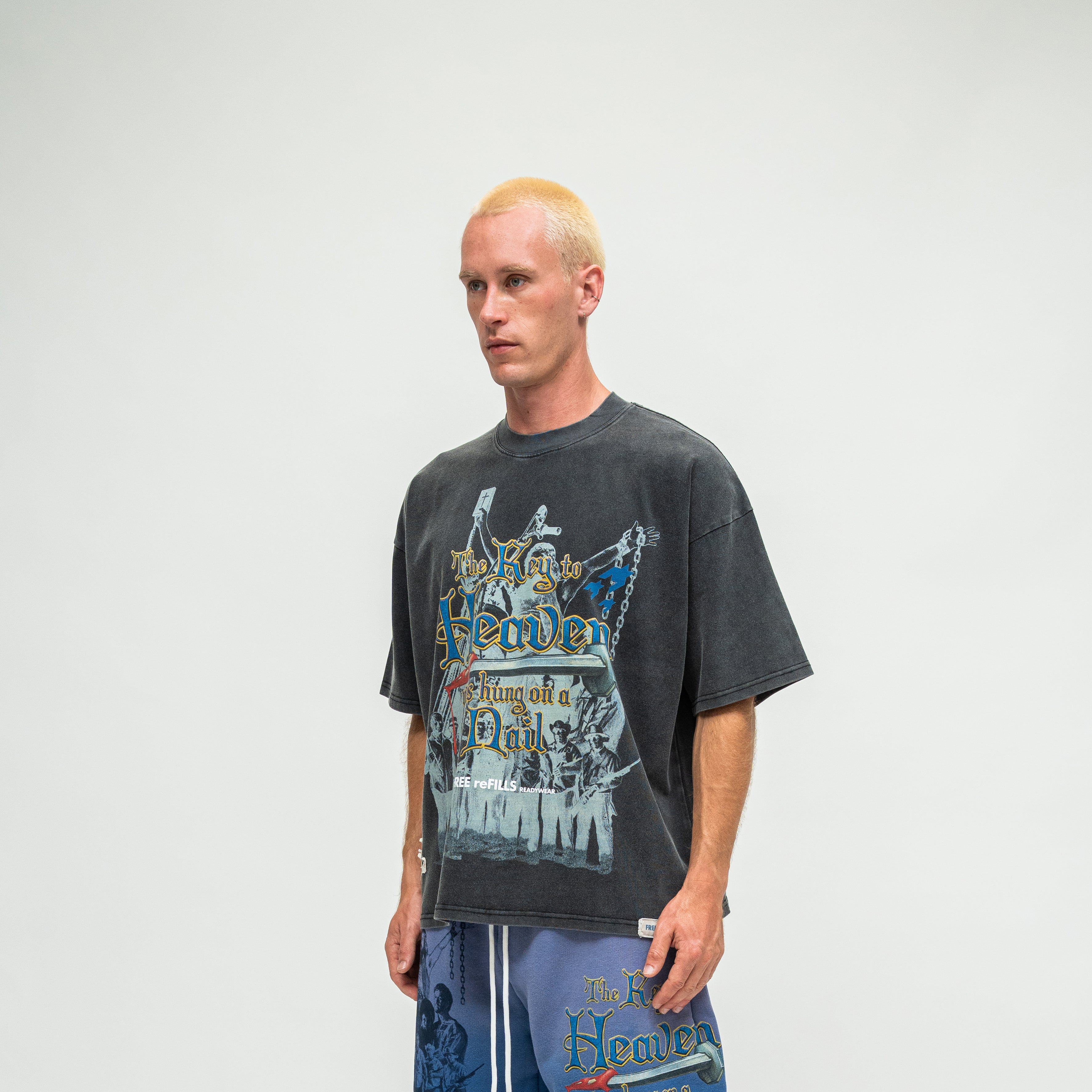 A man with bleached blonde hair wearing a faded black graphic t-shirt and blue pants, standing against a light gray studio background.