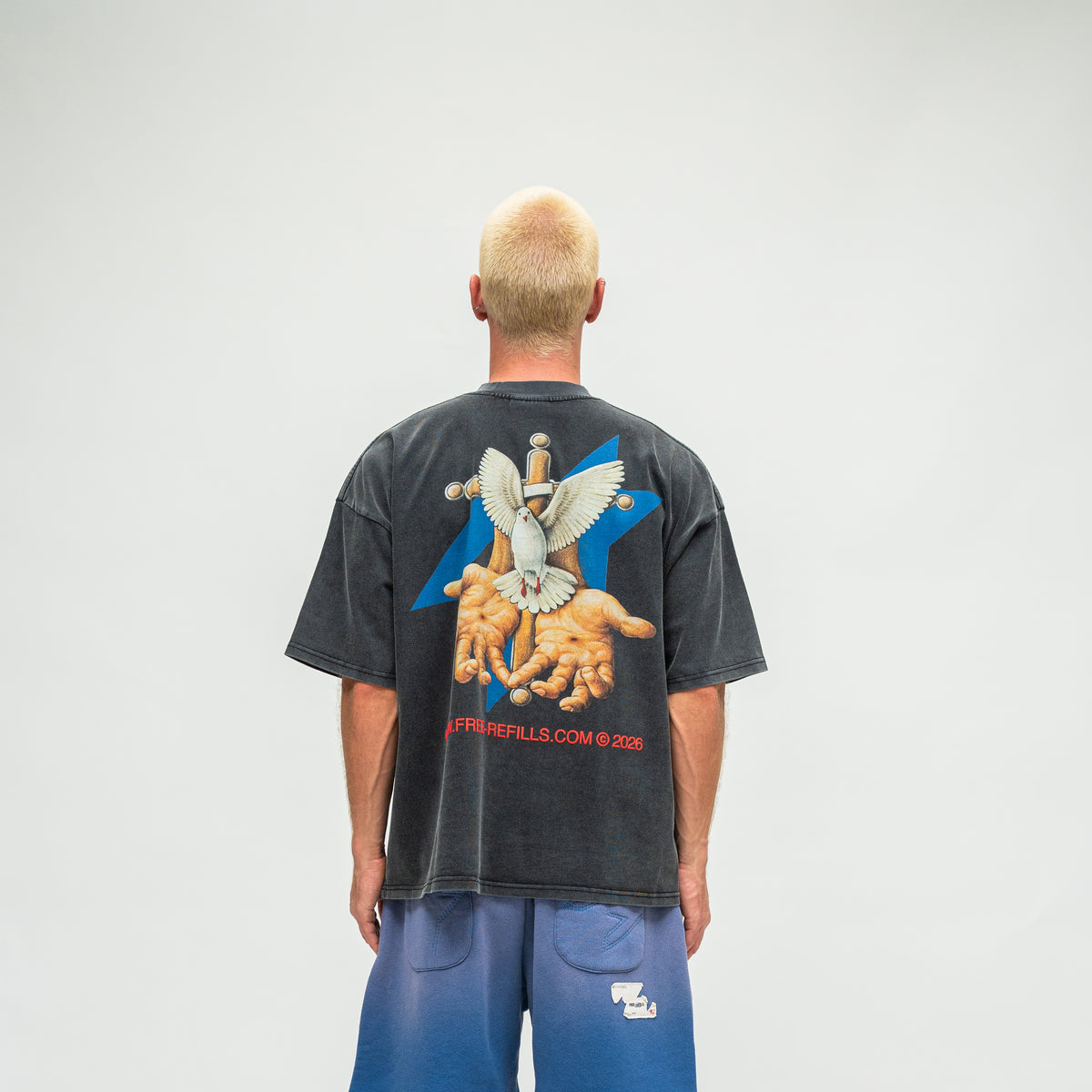 A man viewed from behind wearing a faded black graphic t-shirt and blue sweatpants against a light gray studio background.