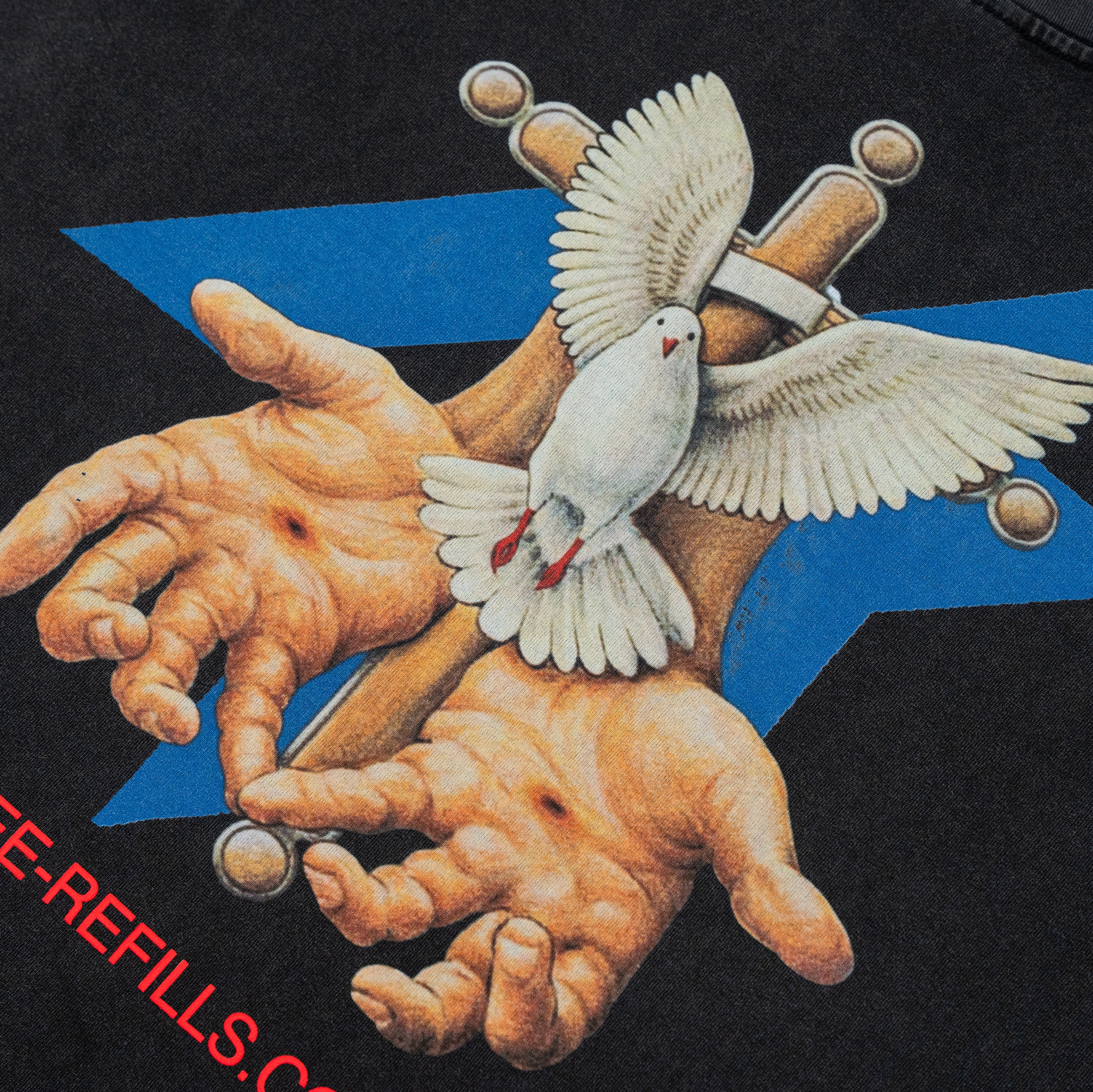 Close-up of a black graphic print featuring two open hands holding crossed metallic objects, with a white dove in flight across the center and a blue geometric shape behind. Partial red text is visible along the bottom left edge.