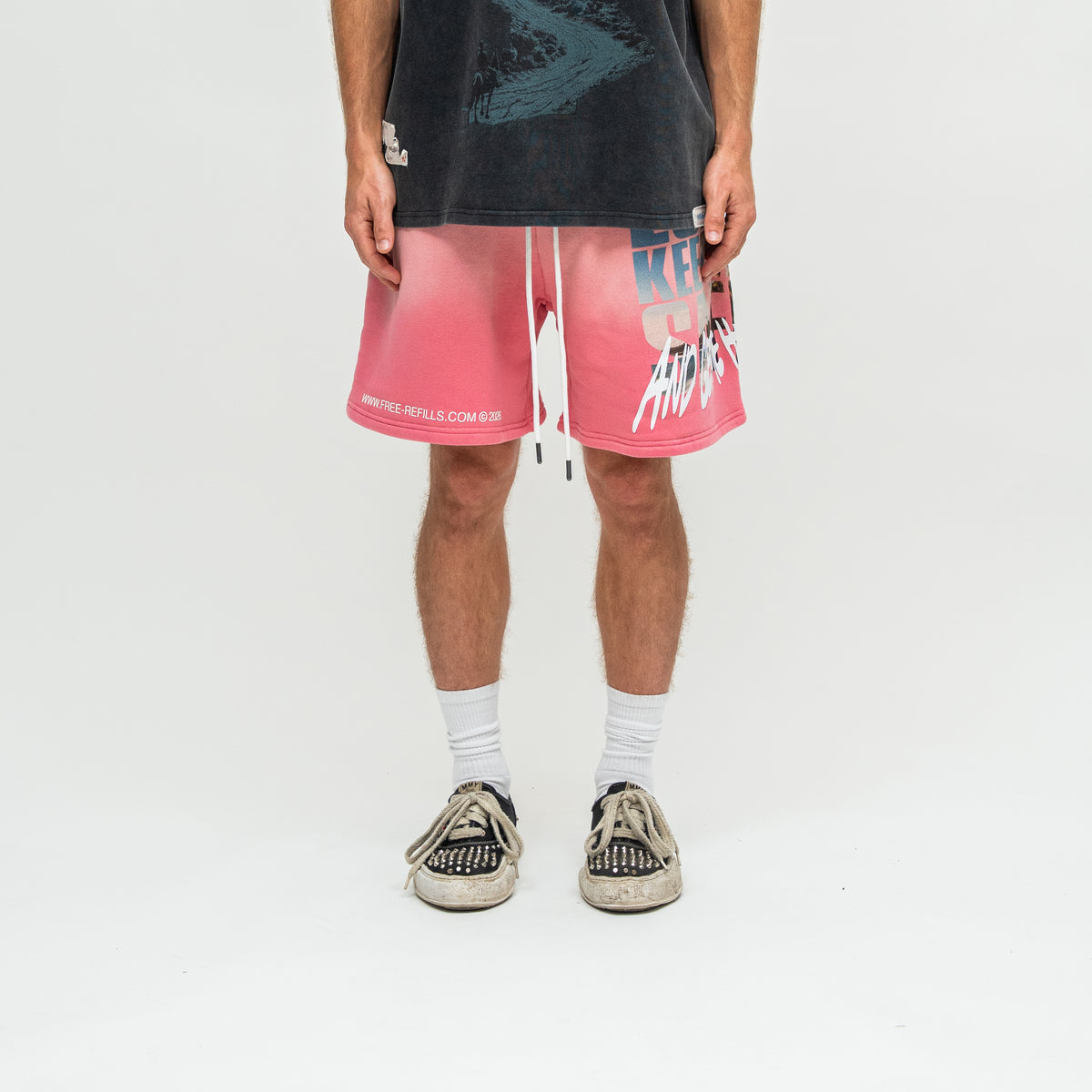 A person standing against a light gray studio background wearing pink gradient drawstring shorts with white graphics and text, a faded black graphic T-shirt, white socks, and black-and-white sneakers.