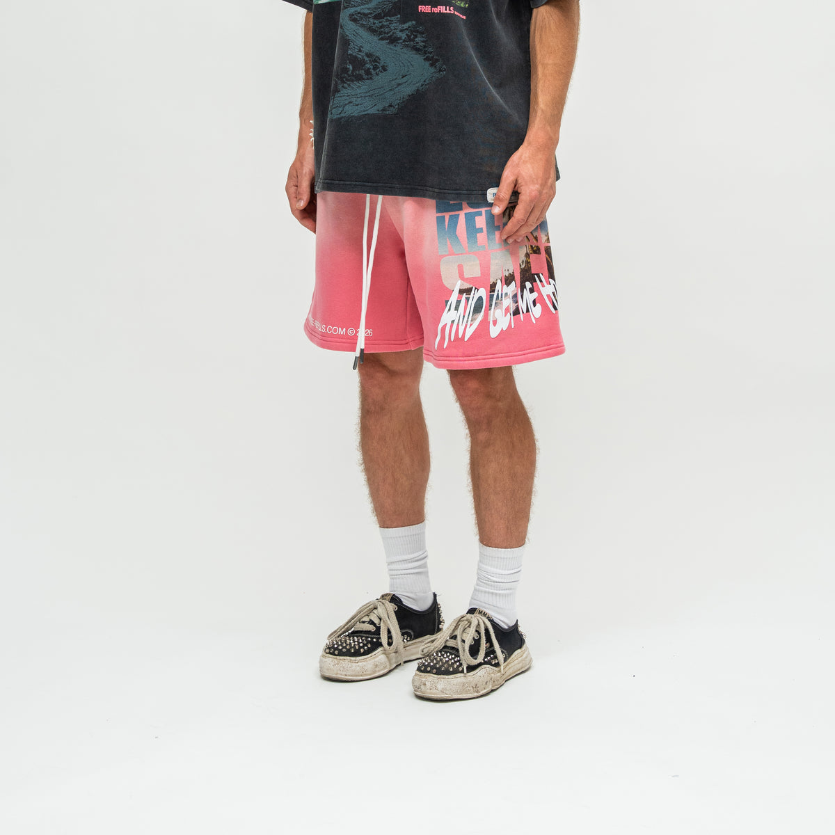 A person wearing pink graphic drawstring shorts, a dark graphic t-shirt, white socks, and black-and-white sneakers, standing against a plain white studio background.