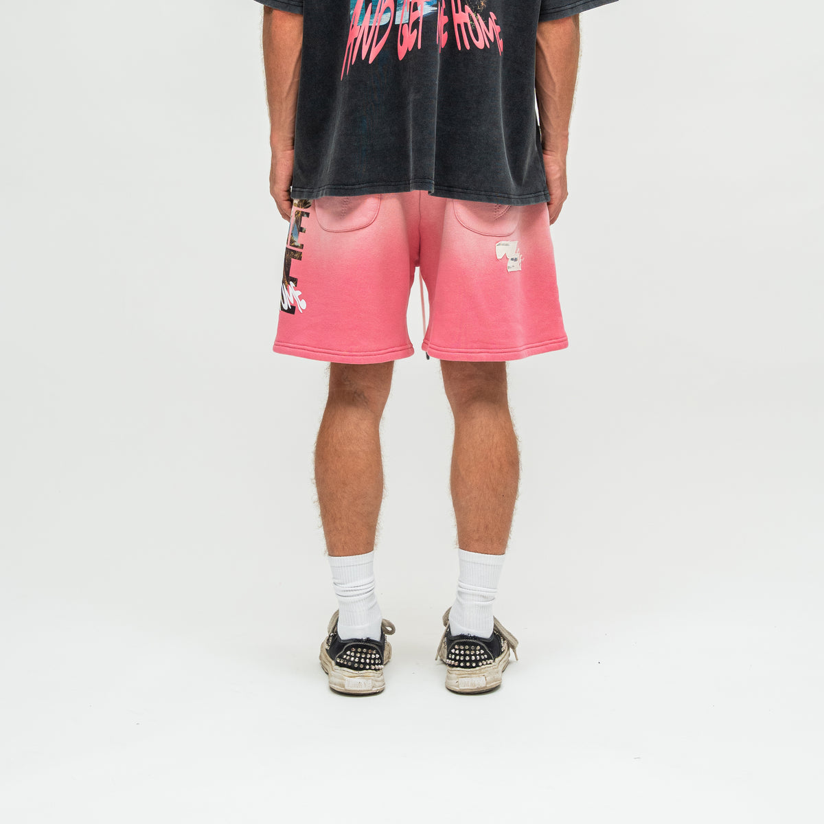 A person shown from the back wearing pink gradient sweat shorts with graphic details, a faded black graphic t-shirt, white socks, and black studded sneakers on a white studio background.