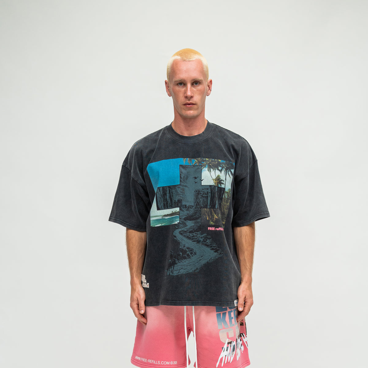 A man modeling an oversized black graphic t-shirt with a blue and green scenic print, paired with pink and white gradient board shorts, against a plain light gray background.