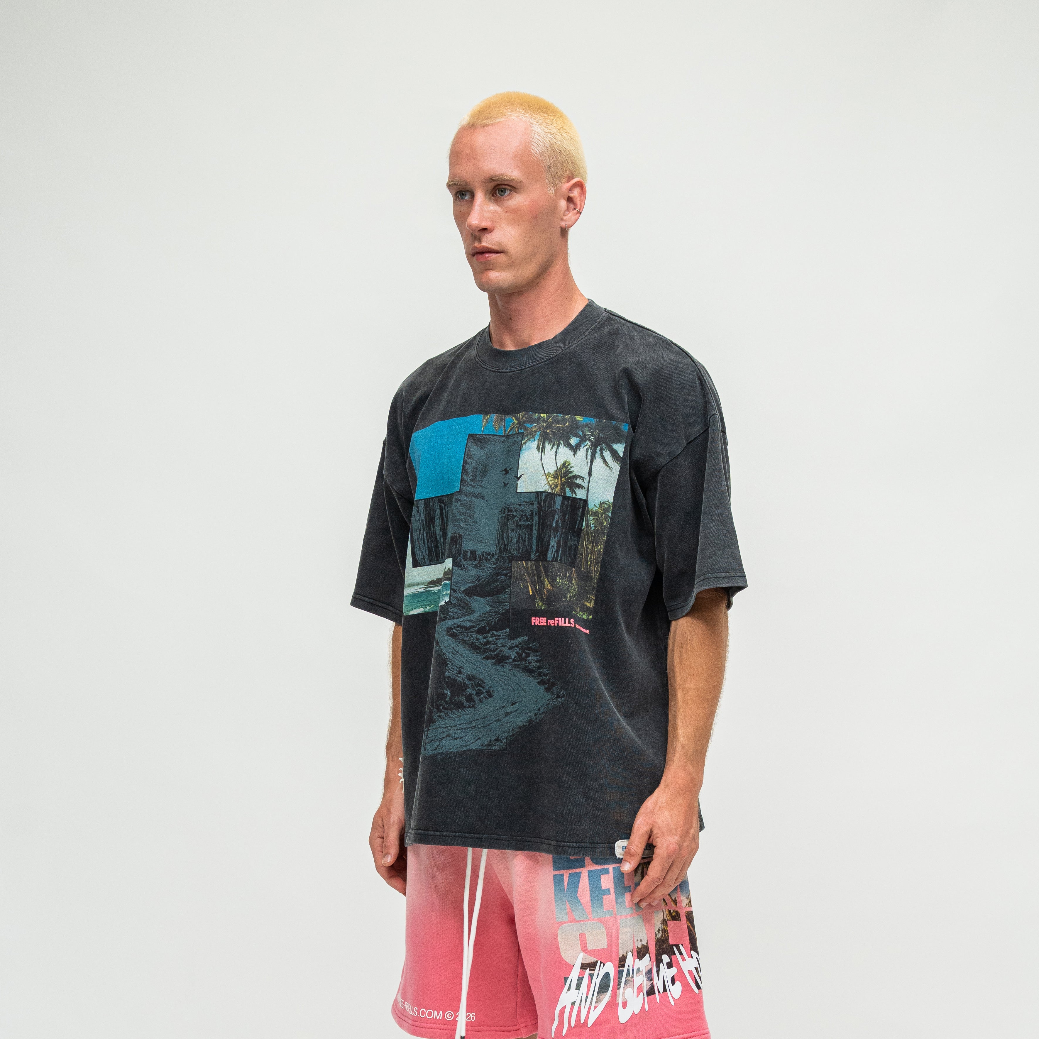 A man with bleached blonde hair stands against a light gray studio background wearing a faded black graphic t-shirt and pink gradient board shorts with white text.