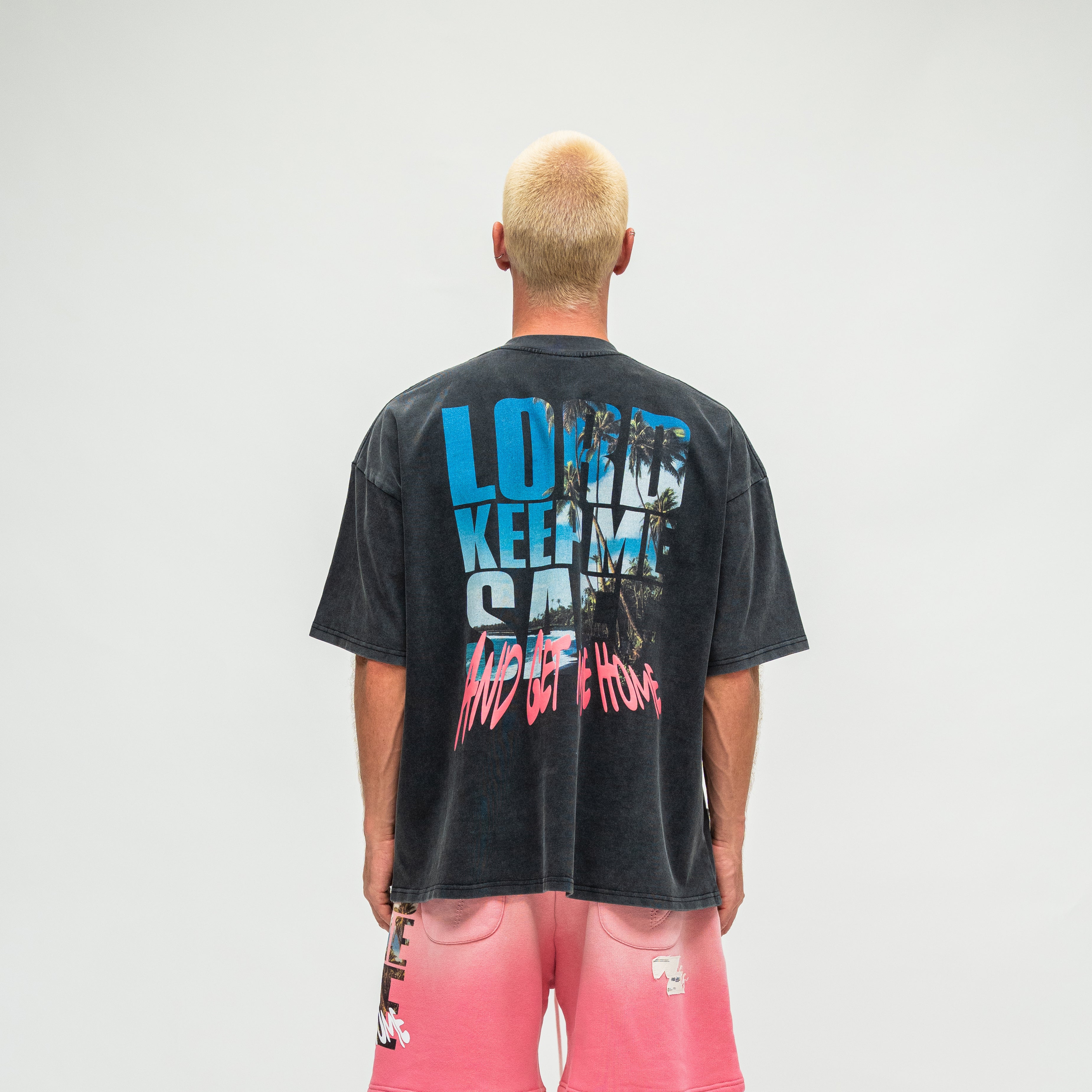 A man viewed from behind wearing a faded black graphic t-shirt with blue and pink text on the back, paired with pink shorts, against a plain light gray background.