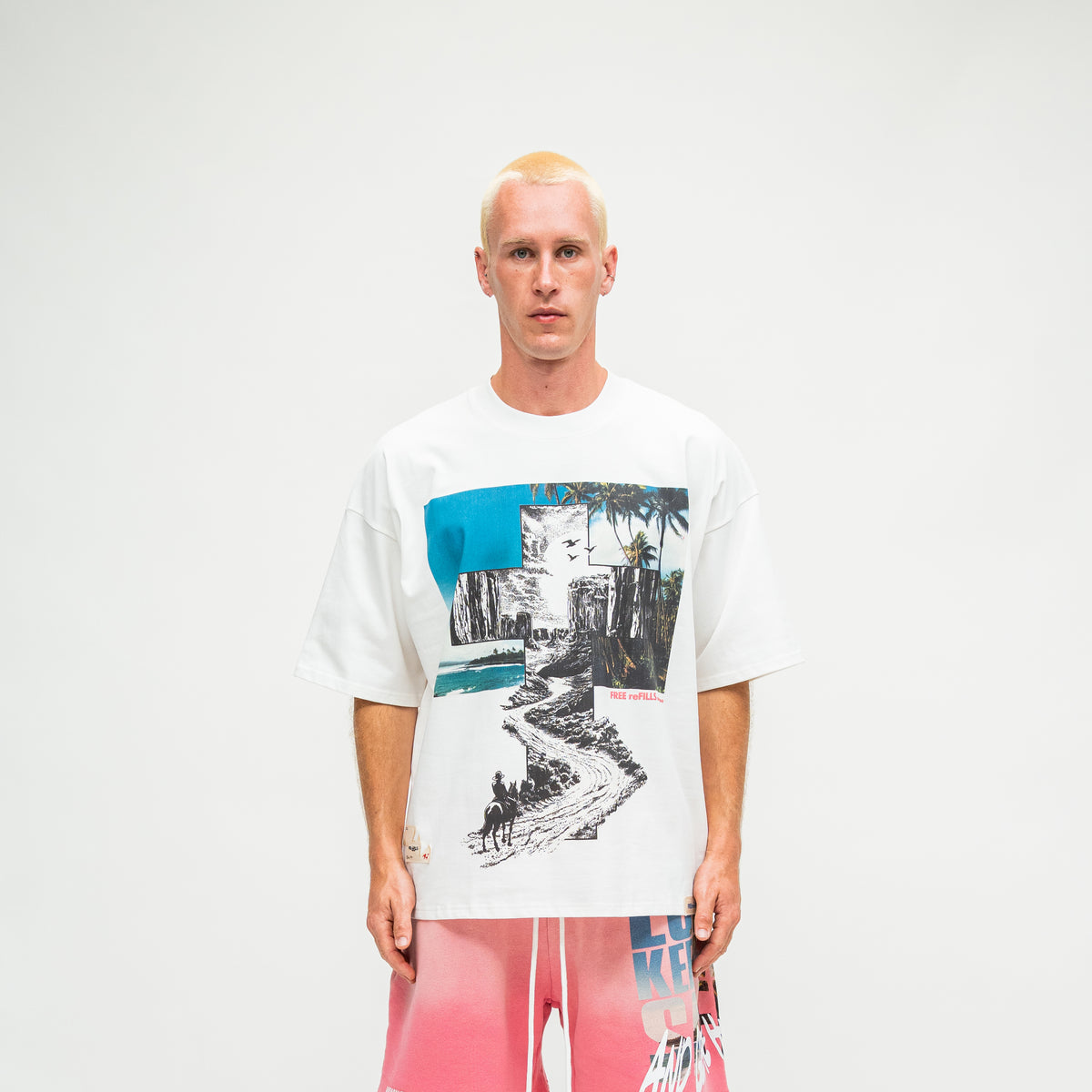 A man wearing a white graphic t-shirt with a colorful collage print and pink gradient shorts, standing against a plain white studio background.