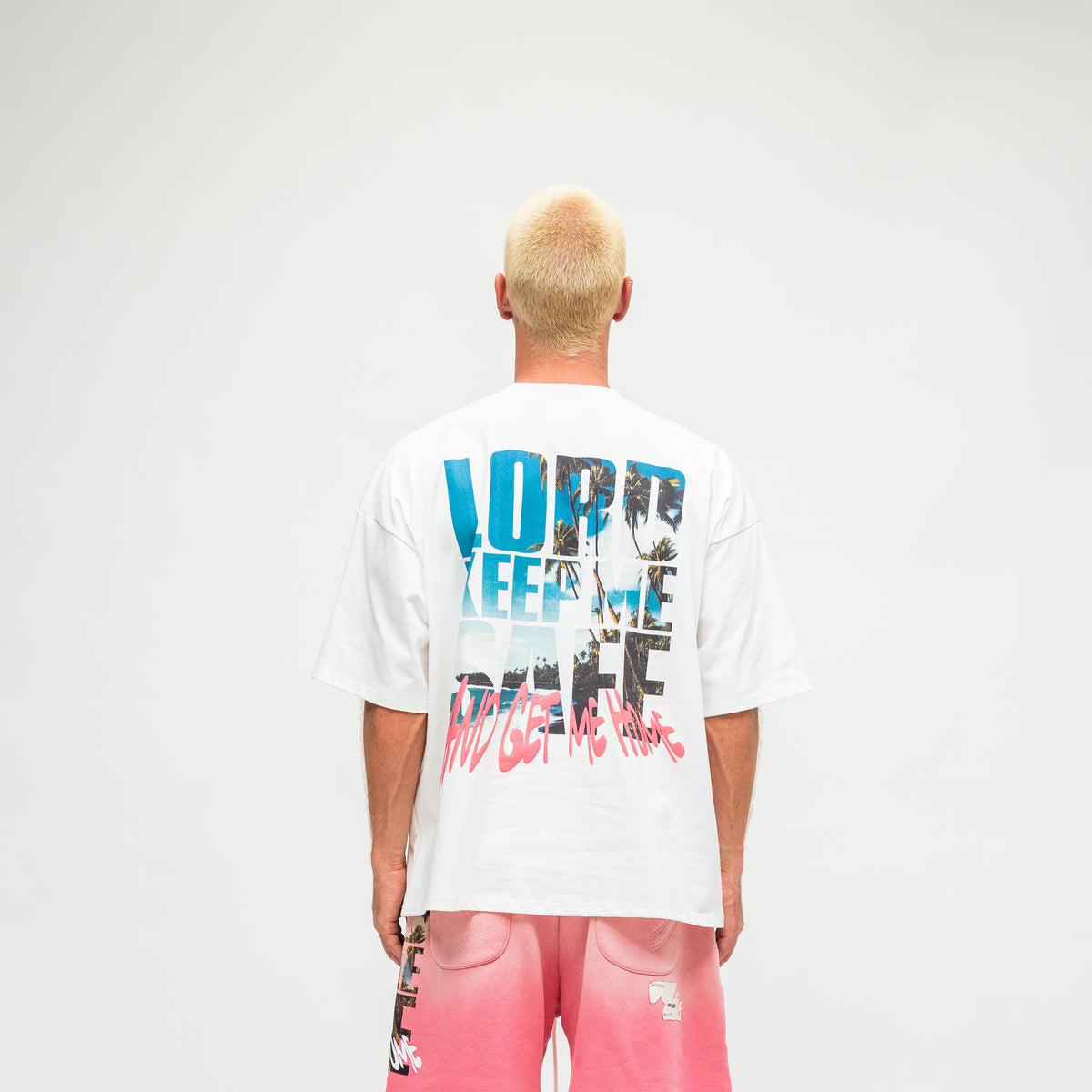 A man viewed from behind wearing a white oversized t-shirt with a large multicolor graphic print on the back, paired with pink shorts, against a plain light gray background.