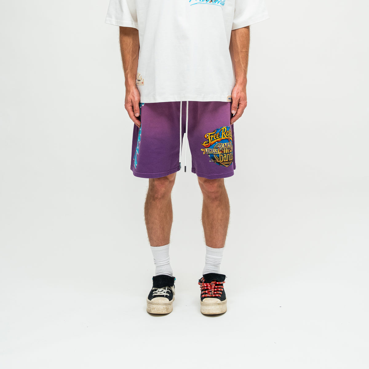 A model from the waist down wearing purple gradient athletic shorts with a yellow and blue graphic on the left leg, styled with a white t-shirt, white socks, and black sneakers with red laces against a white studio background.