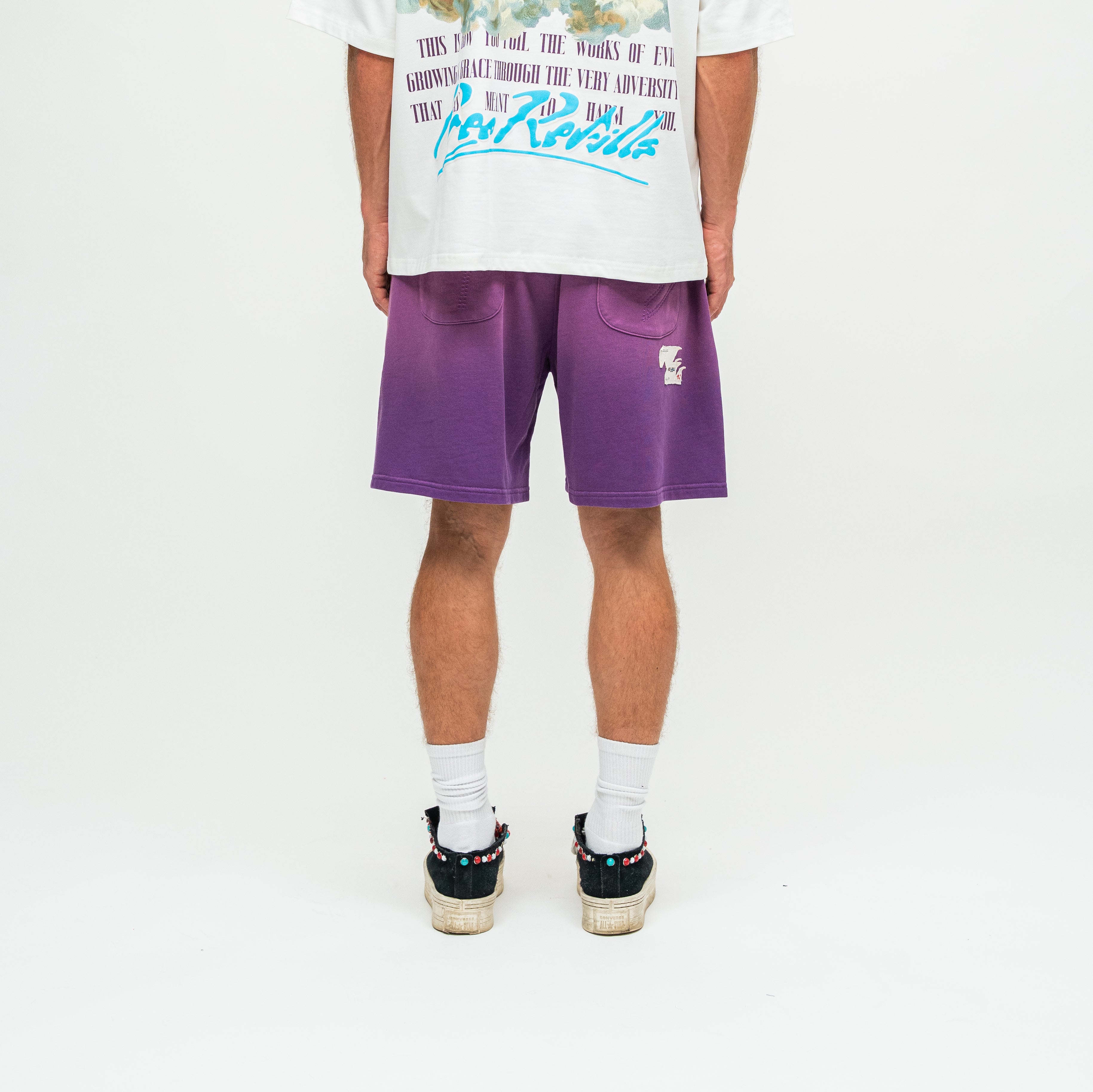 A person shown from the back wearing purple shorts, a white graphic T-shirt, white socks, and black sneakers with colorful details, photographed against a clean white studio background.