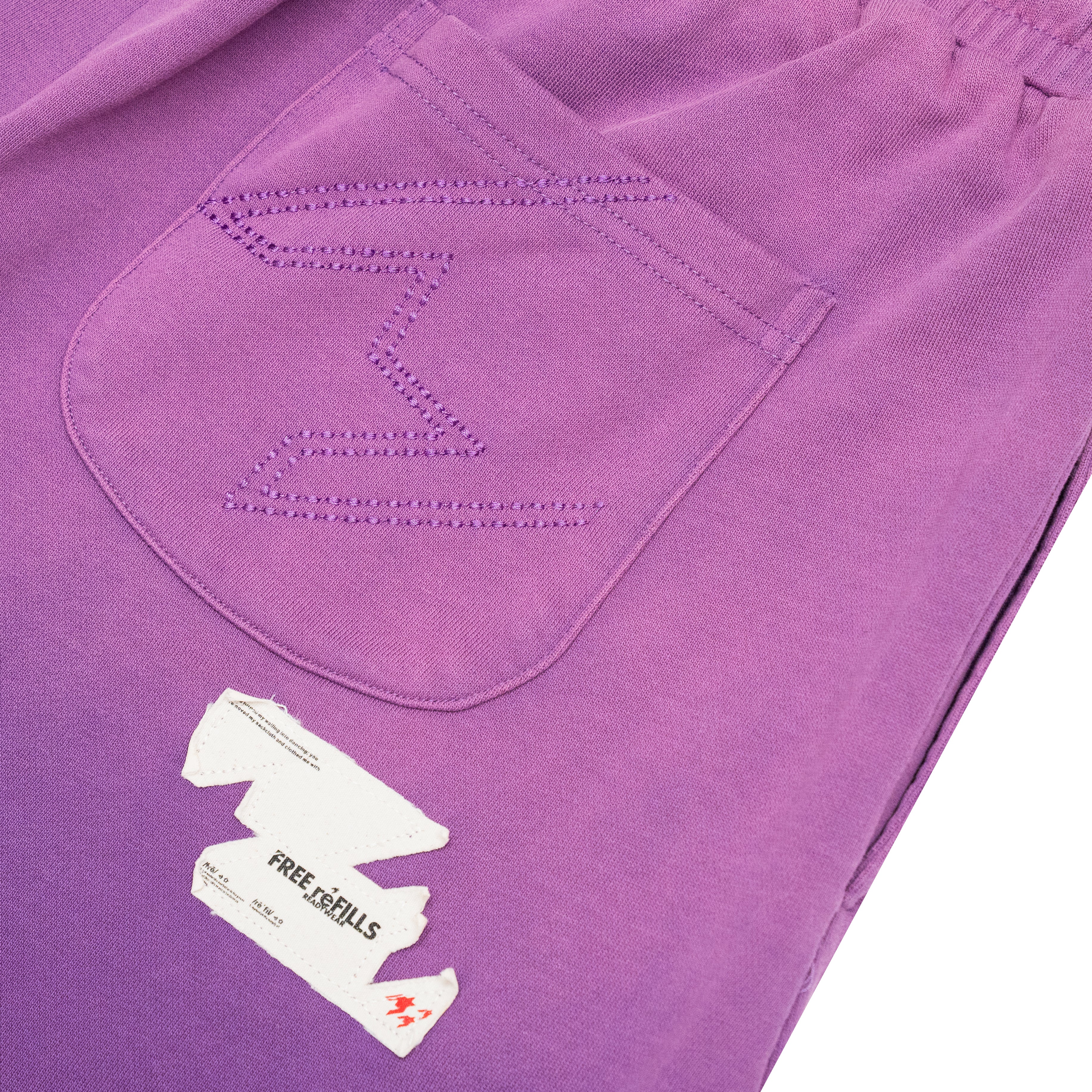Close-up of purple corduroy trousers showing a stitched pocket with a zigzag pattern and a white paper tag that reads FREE REFILLS.