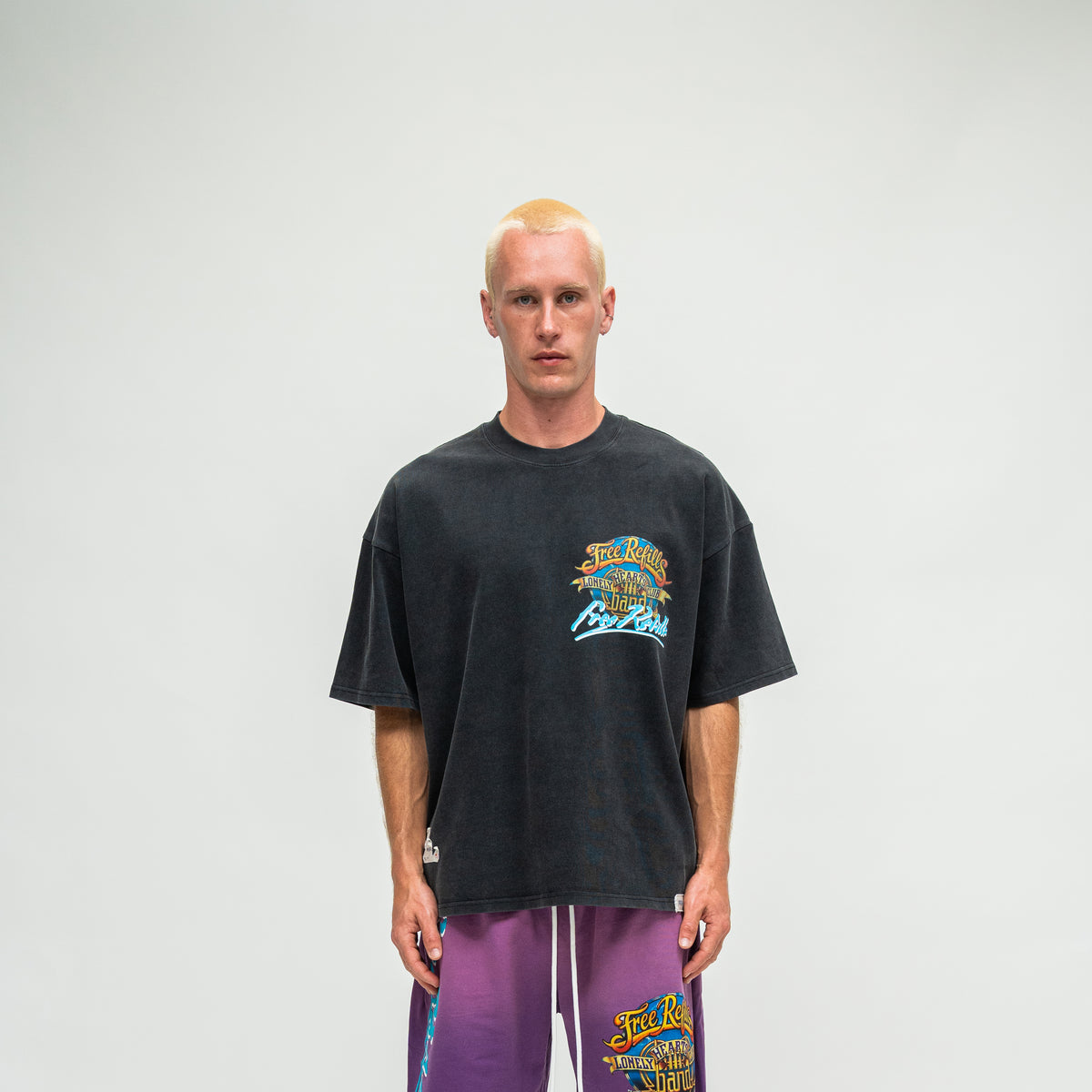 A man wearing a faded black graphic t-shirt and purple sweatpants, standing against a light gray studio background.