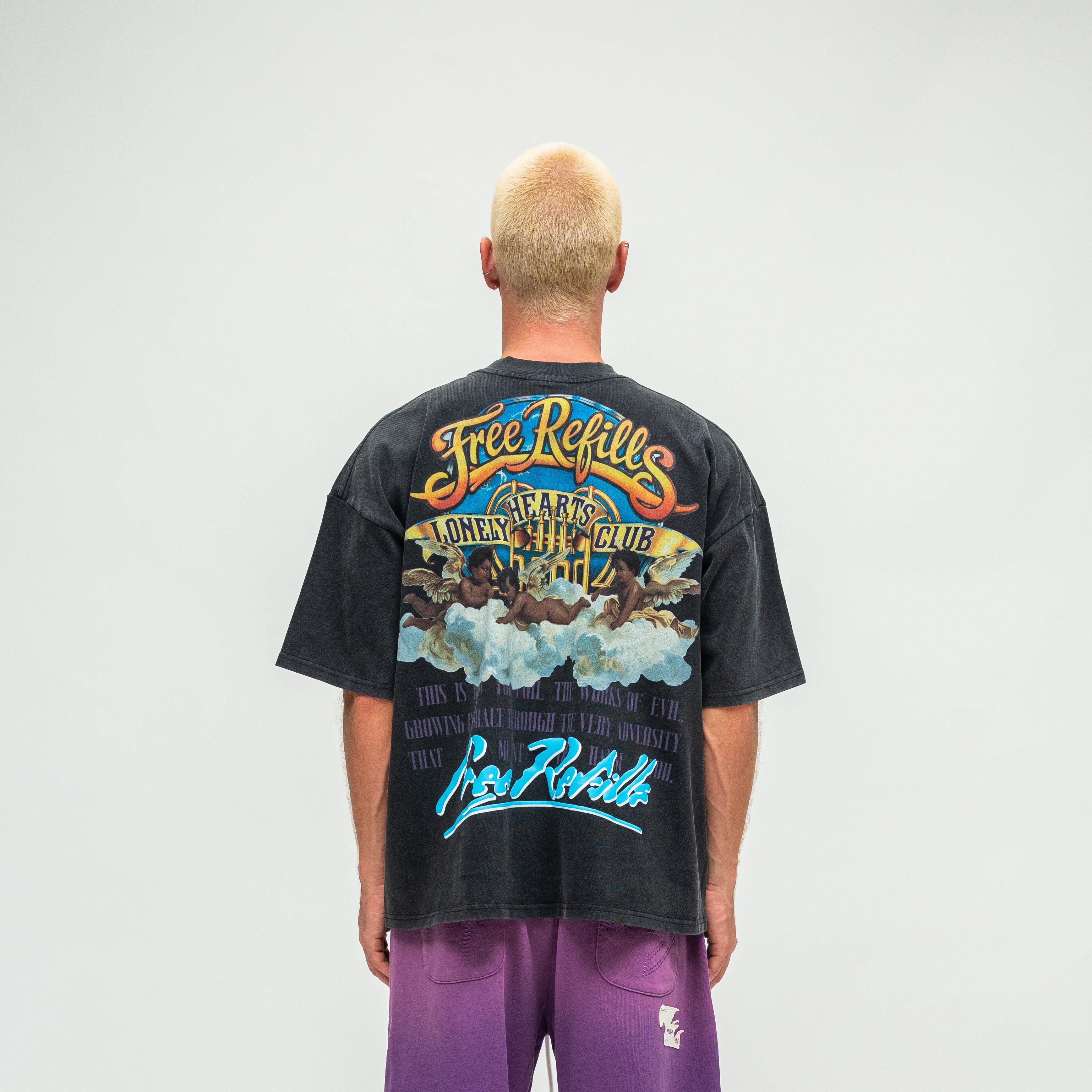 A man viewed from behind wearing a faded black graphic t-shirt with a colorful 'Free Refills' print and purple pants, set against a plain light gray background.